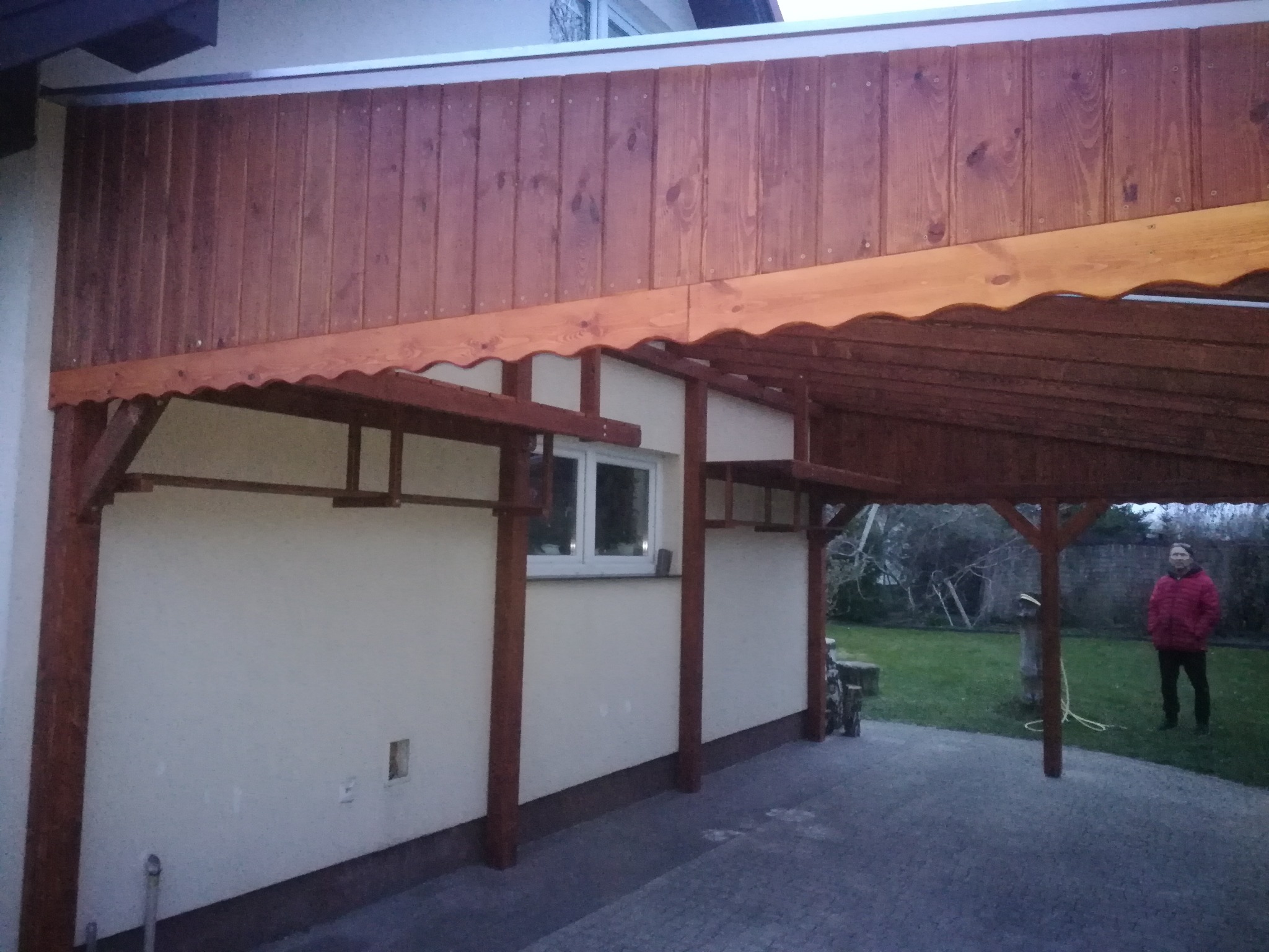Zadaszenie drewniane z falistym wykończeniem, wsparte na słupach, przylegające do budynku z oknem, na zewnątrz widoczna osoba w czerwonej kurtce na trawniku.
