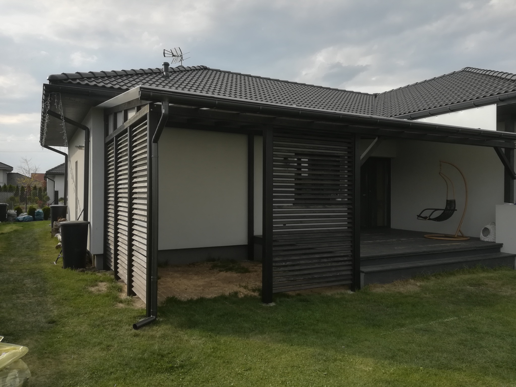 Nowoczesna konstrukcja z drewnianych paneli osłaniająca taras przy domu z ciemną dachówką, widoczny fragment ogrodu i wiszące krzesło.