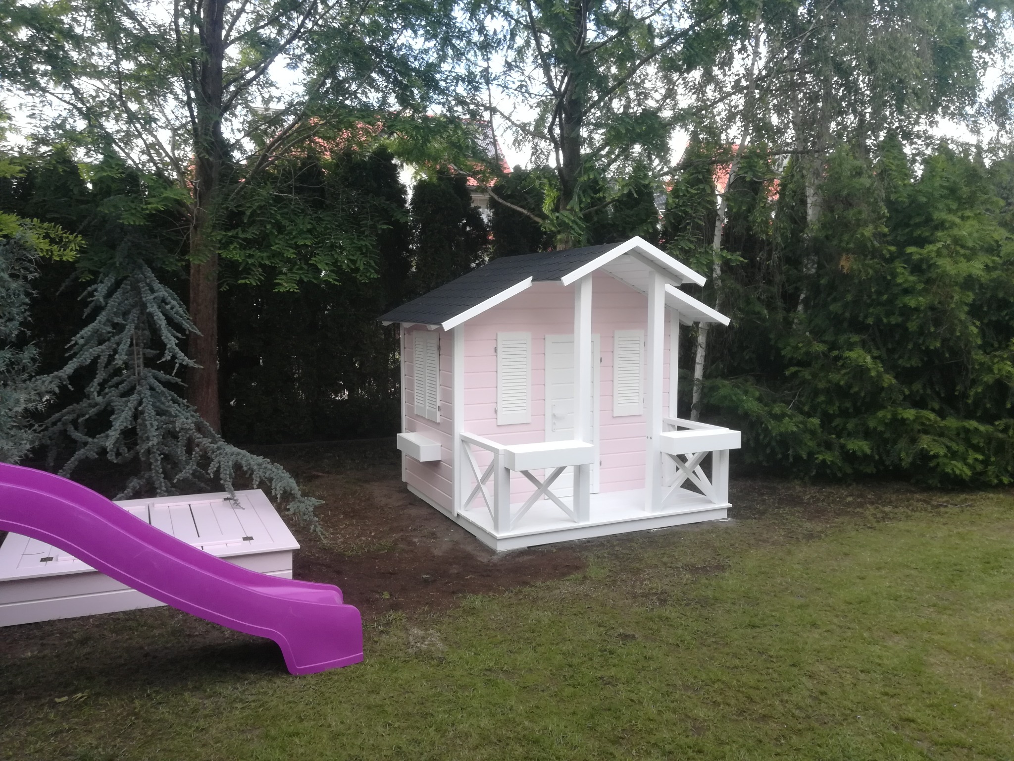 Różowy drewniany domek dla dzieci z białymi okiennicami i balustradą, z fioletową zjeżdżalnią na trawniku, otoczony zielenią drzew i krzewów.