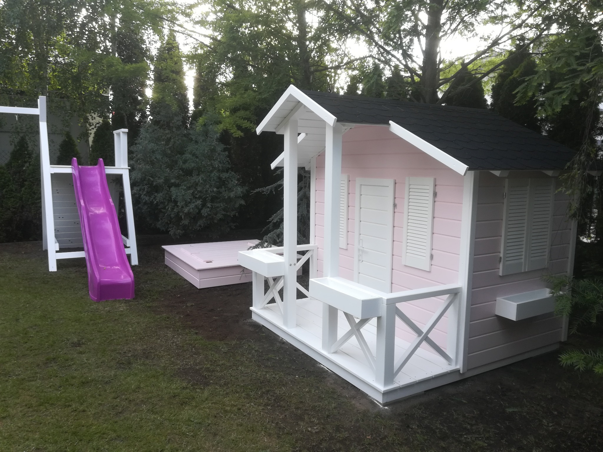 Różowy domek zabaw dla dzieci z białymi elementami, czarnym dachem, werandą, piaskownicą i fioletową zjeżdżalnią na trawniku w ogrodzie.