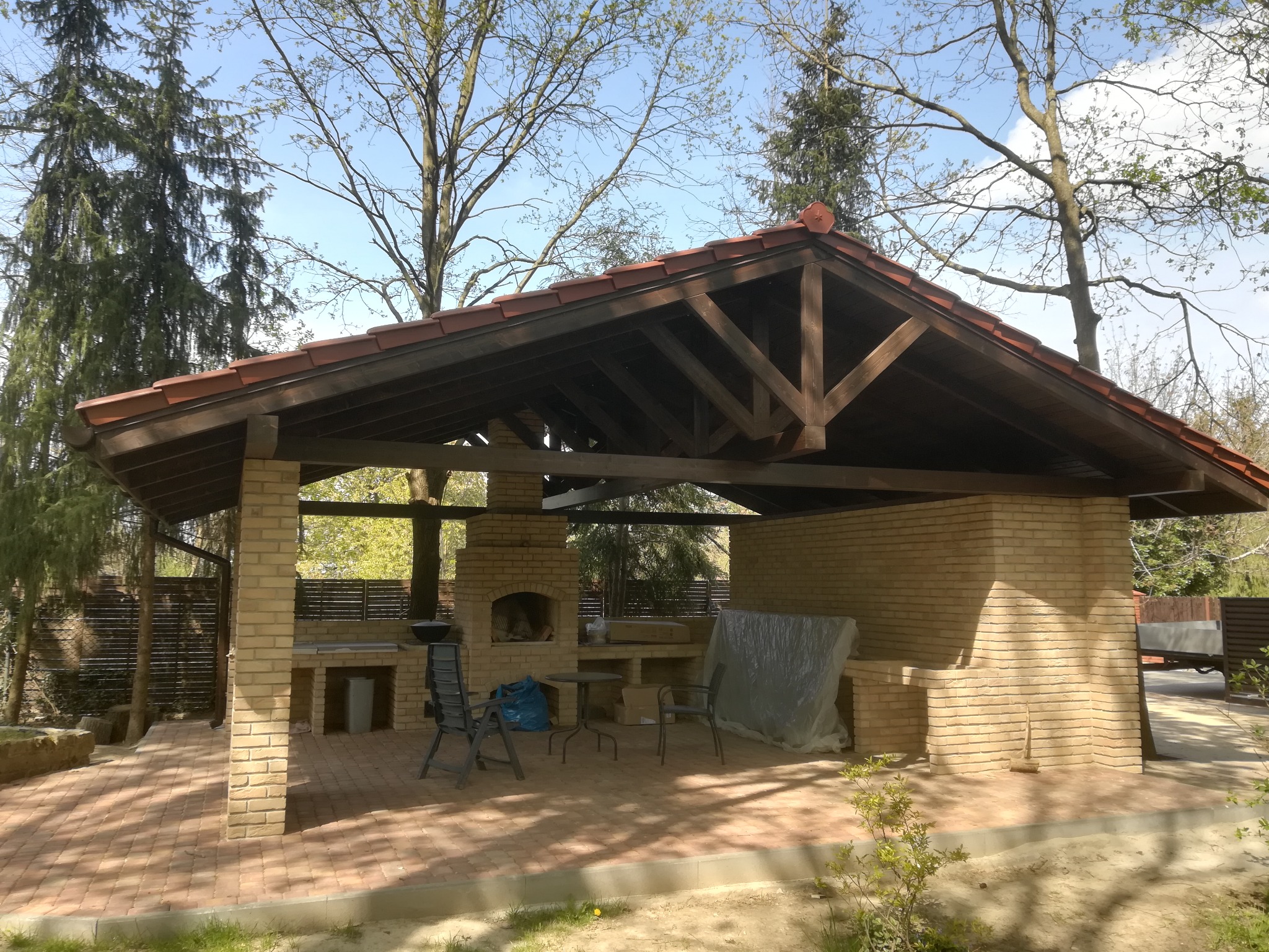 Murowana altana z cegły klinkierowej z kominkiem, zadaszona konstrukcją drewnianą i czerwoną dachówką, na wybrukowanym tarasie.