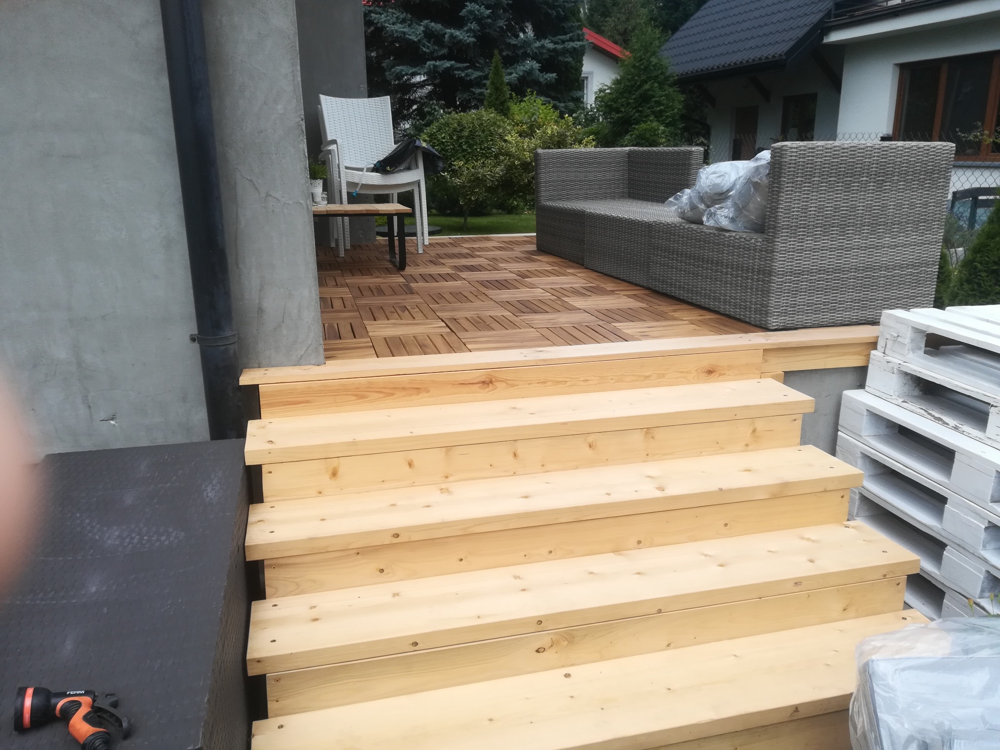 Fragment tarasu z drewnianą podłogą z płytek i schodami z surowego drewna, na tle domu z szarą elewacją i meblami ogrodowymi, widoczne narzędzie i palety