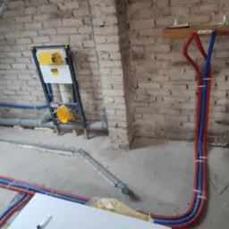 Wnętrze w trakcie prac instalacyjnych w surowym stanie: widoczny stelaż podtynkowy WC marki Viega, rury CO i wodne prowadzone po podłodze i na ścianie z cegły, szare rury kanalizacyjne z pianką...