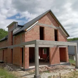 Dwukondygnacyjny budynek w stanie surowym otwartym, z czerwonej cegły, z częściowo wykonanym dachem z blachy trapezowej, widoczne betonowe słupy podtrzymujące zadaszenie tarasu, w tle zieleń...