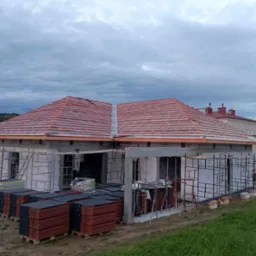 Niedokończony dom z szarej cegły z częściowo położonym, czerwonym dachem, widoczne rusztowania i palety z dachówką.