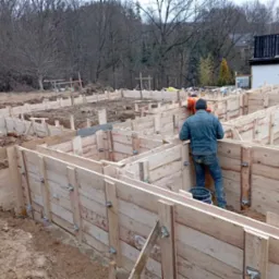 Szczegółowy widok szalunków fundamentowych z widocznymi pracownikami w trakcie montażu, z fragmentem zabudowy w tle.