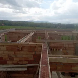 Wznoszenie ścian domu z czerwonej cegły, widoczne szalunki i drabiny, w tle krajobraz z polami i lasami pod pochmurnym niebem.