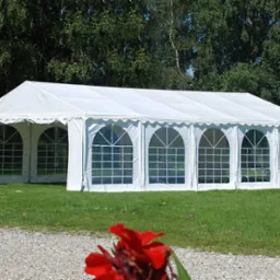 Rozłożony biały namiot imprezowy z oknami w kształcie łuków, widoczny na trawniku z czerwonymi kwiatami w rogu kadru.