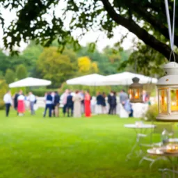 Ogr&oacute;d z białymi parasolami i grupą ludzi w tle, w pierwszym planie lampion zawieszony na drzewie, stoliki z krzesłami.