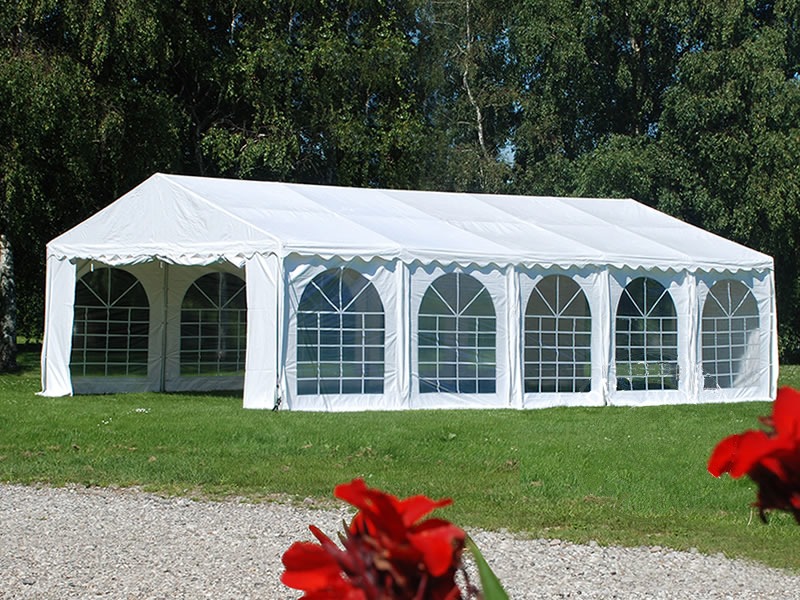 Rozłożony biały namiot imprezowy z oknami w kształcie łuków, widoczny na trawniku z czerwonymi kwiatami w rogu kadru.