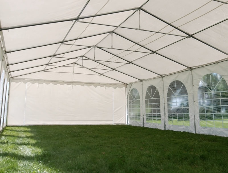 Wnętrze białego namiotu imprezowego z widokiem na zielony trawnik, przez okna widać drzewa.