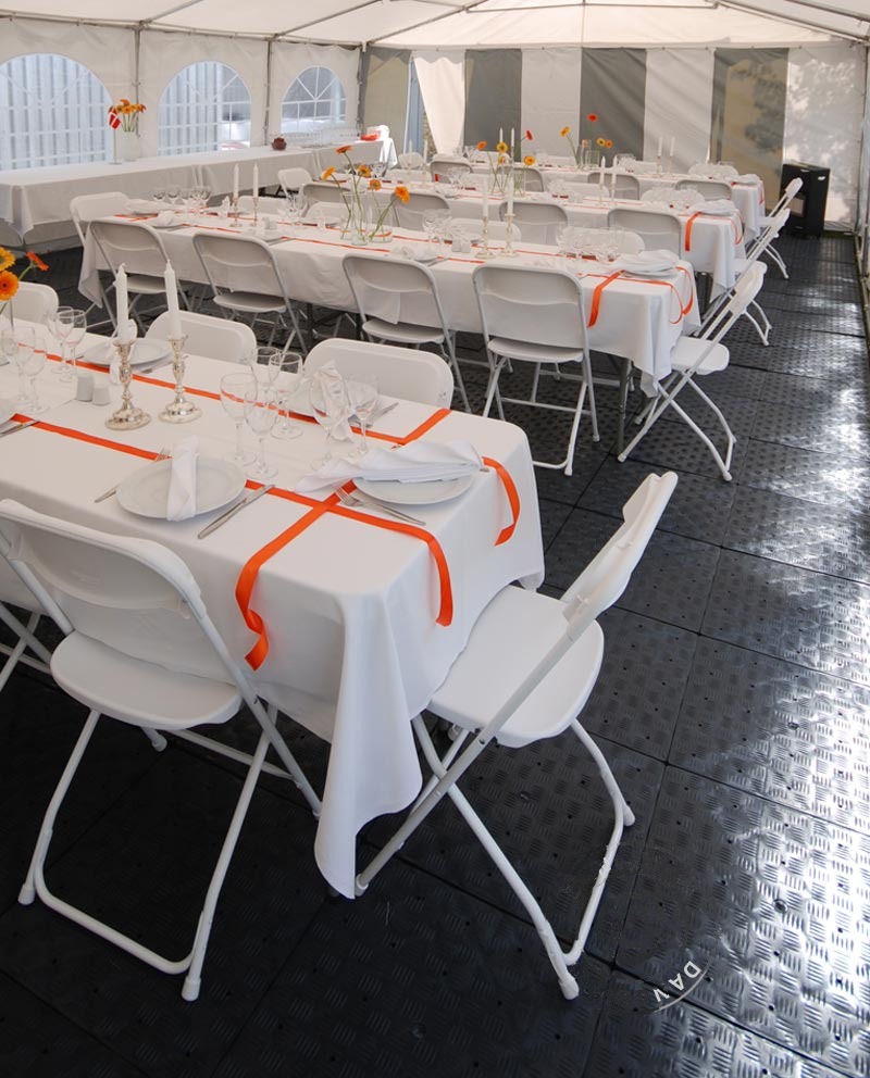 Uroczysta aranżacja stołów w białym namiocie: białe obrusy, pomarańczowe wstążki, proste białe krzesła, zastawa stołowa, świeczniki i kwiaty, czarna podłoga z fakturą.