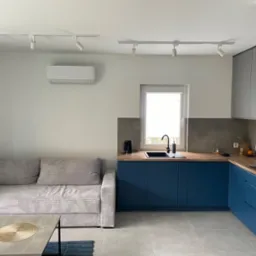 Nowoczesne wnętrze mieszkania z klimatyzatorem na ścianie, niebieską zabudową kuchenną i szarą sofą.