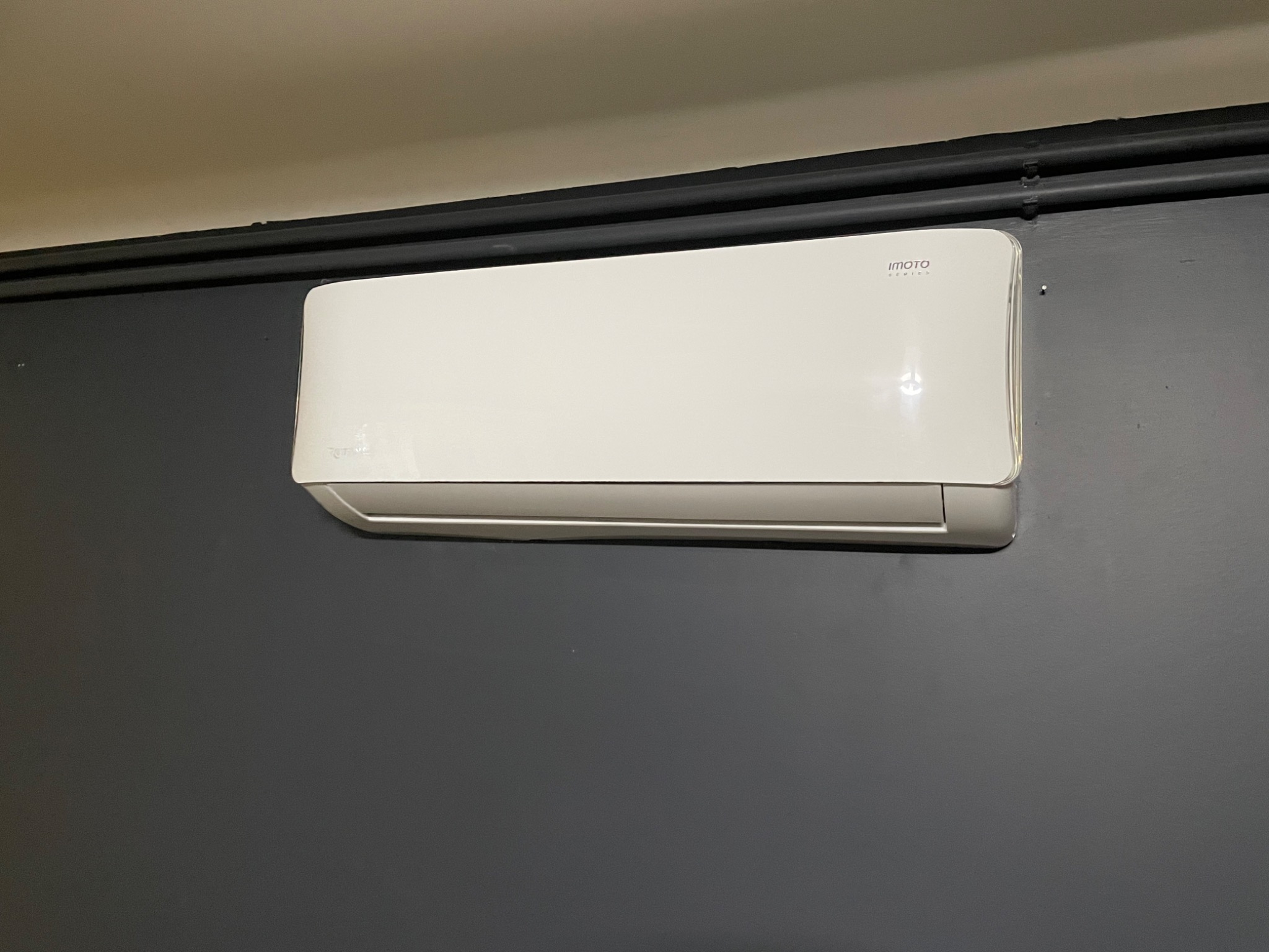 Biały klimatyzator marki Imoto zamontowany na szarej ścianie, widoczna czarna rura instalacyjna pod sufitem.