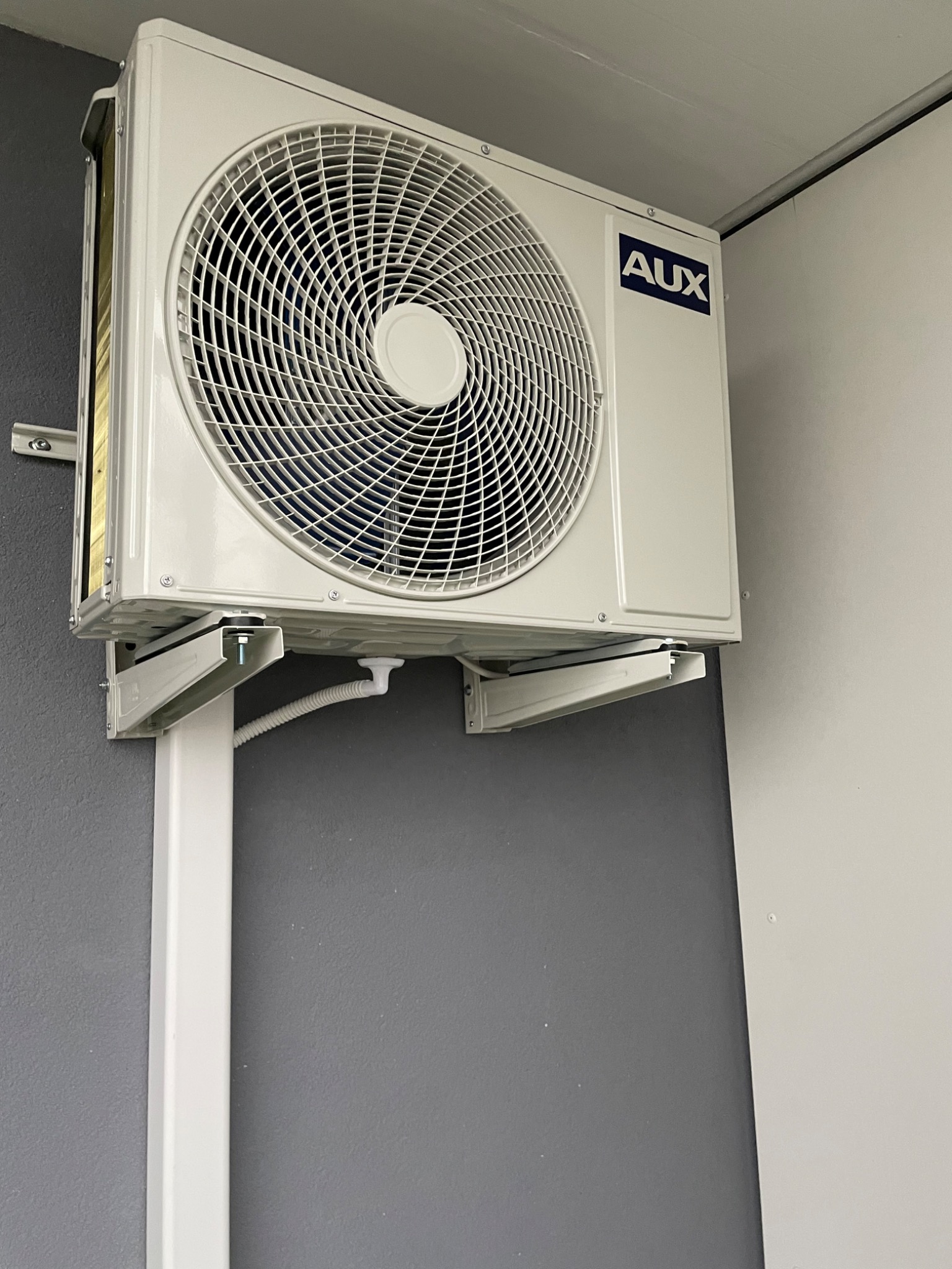 Biała jednostka zewnętrzna klimatyzatora marki AUX zamontowana na wspornikach na szarej ścianie, widoczne białe rury instalacyjne.