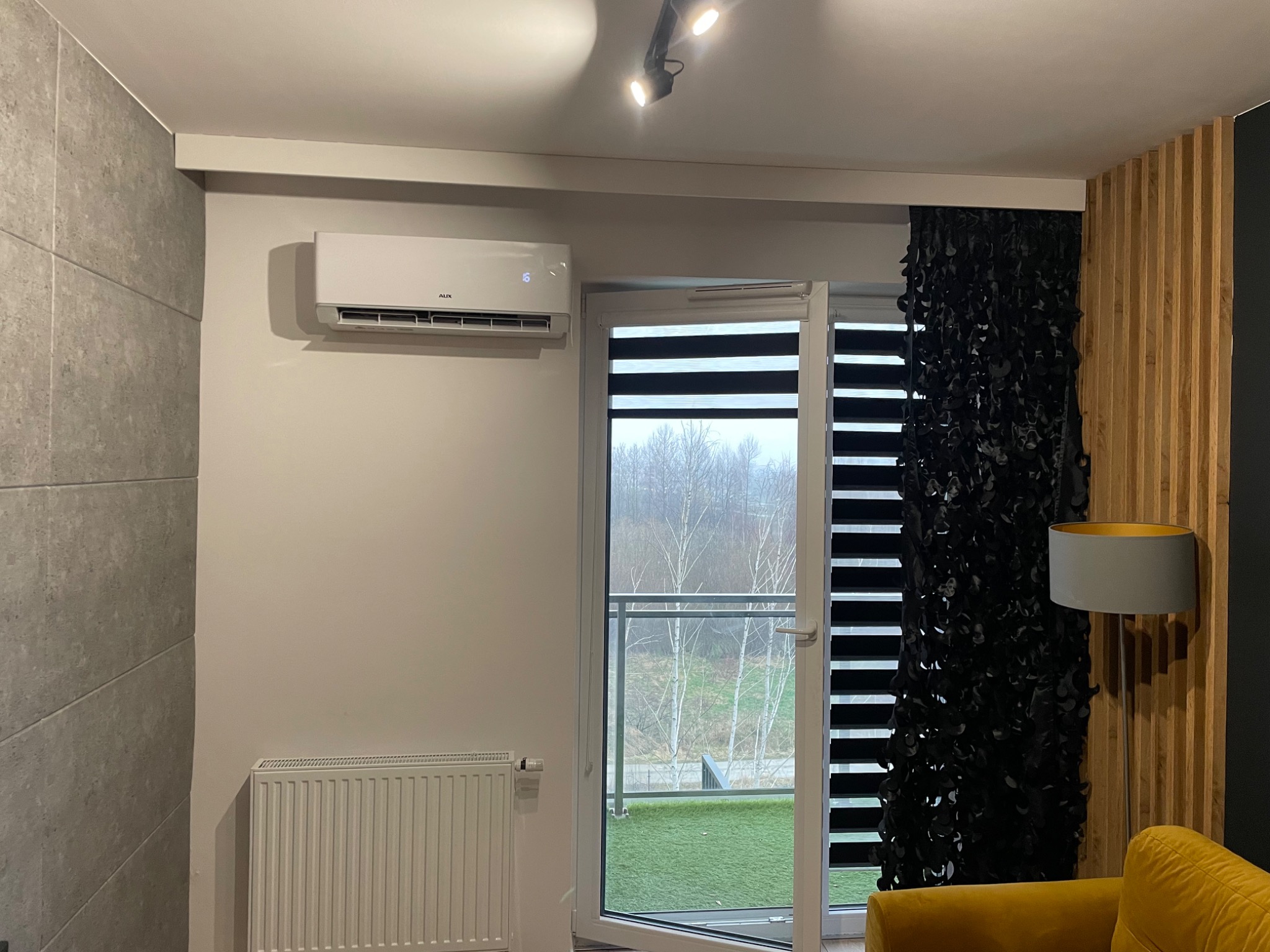 Nowoczesny klimatyzator ścienny marki AUX zamontowany w jasnym salonie z wyjściem na balkon, widoczne żaluzje okienne i dekoracyjna zasłona z czarnych elementów, grzejnik pod oknem.
