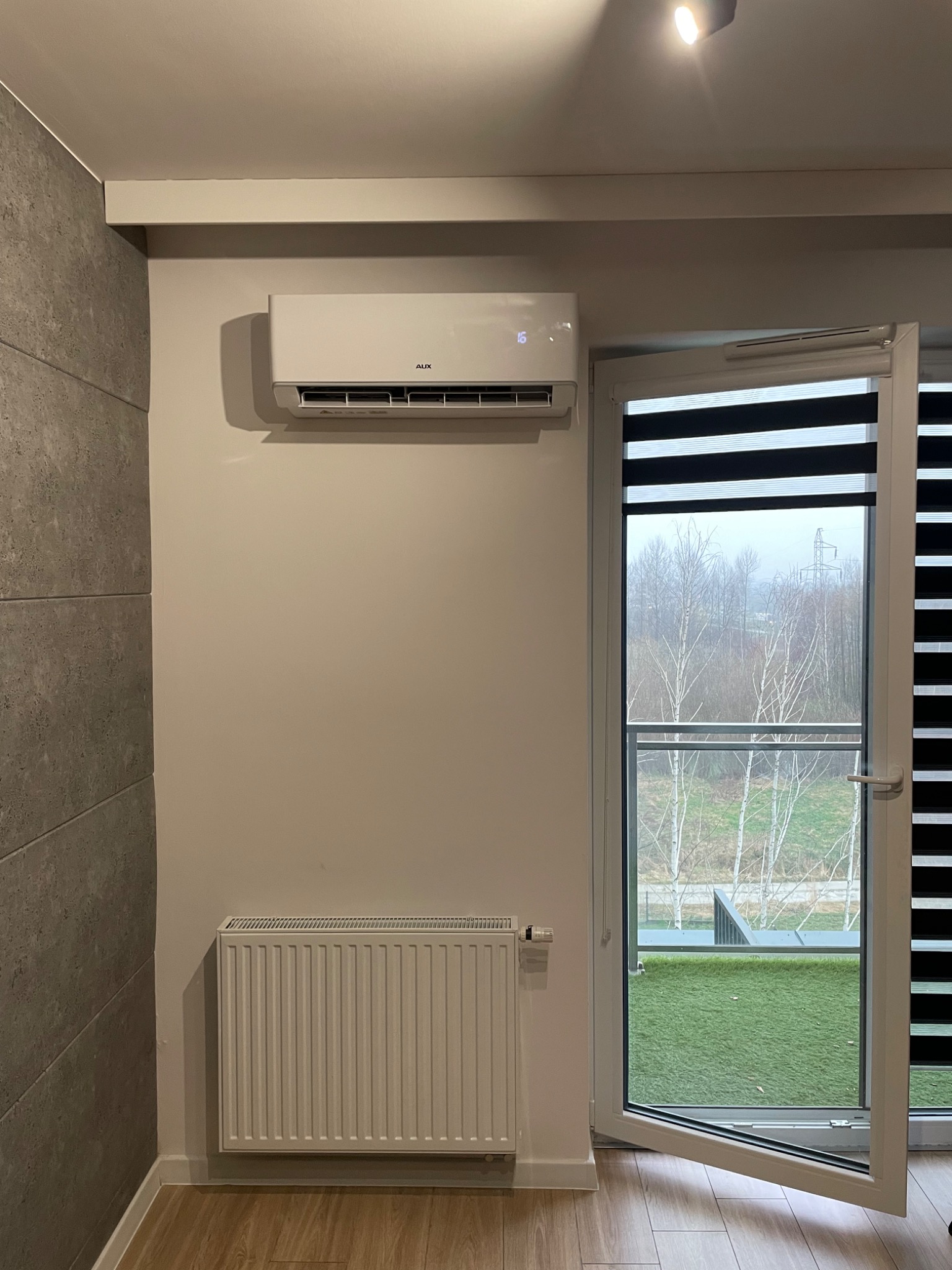 Biały klimatyzator ścienny marki AUX zamontowany nad grzejnikiem w nowoczesnym wnętrzu z szarą ścianą i otwartym oknem balkonowym z widokiem na drzewa i sztuczną trawę.