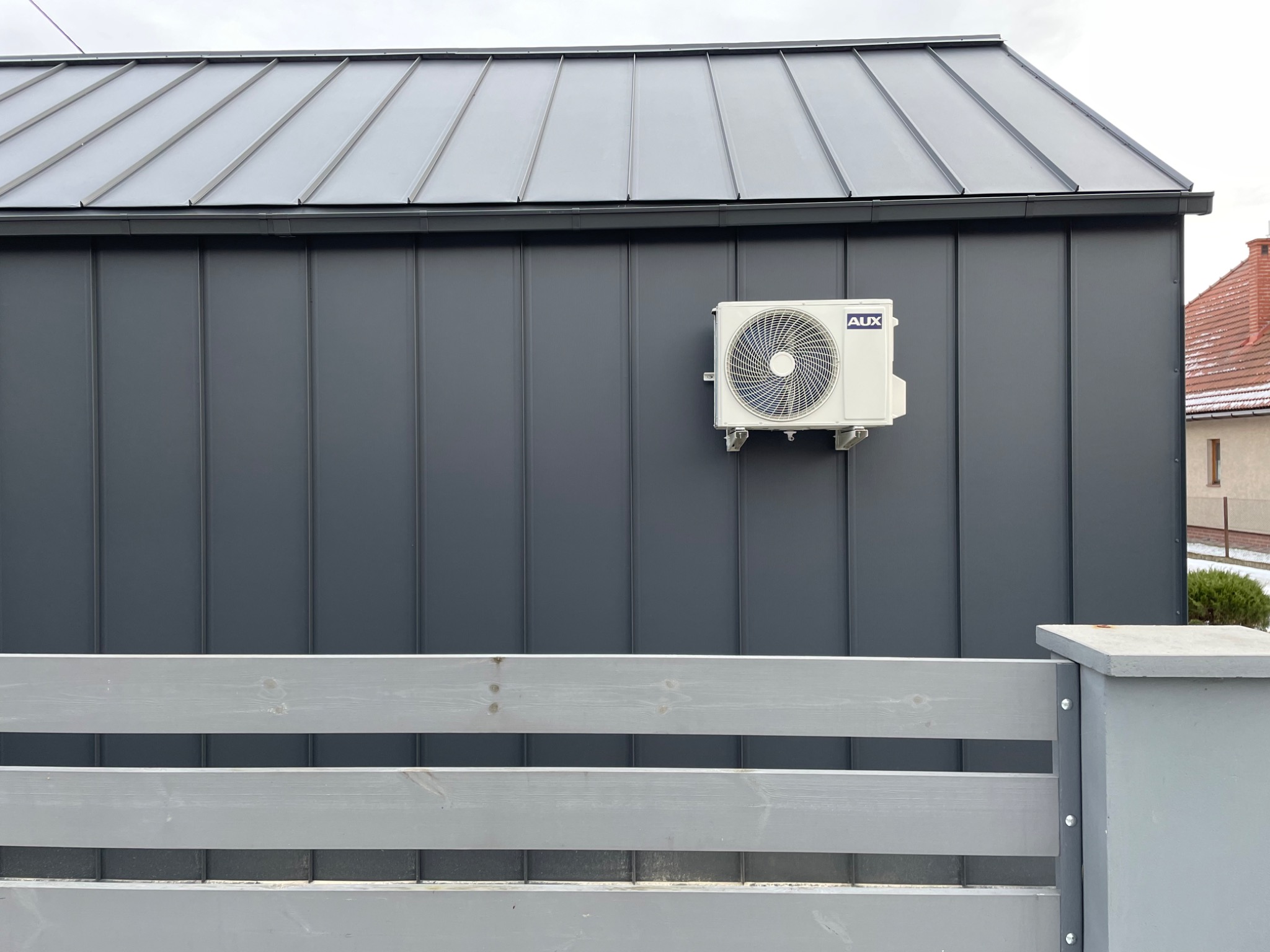 Biała jednostka zewnętrzna klimatyzacji marki AUX zamontowana na szarej, metalowej ścianie budynku z panelami pionowymi i szarym dachem z blachy trapezowej, widoczny fragment szarego ogrodzenia.