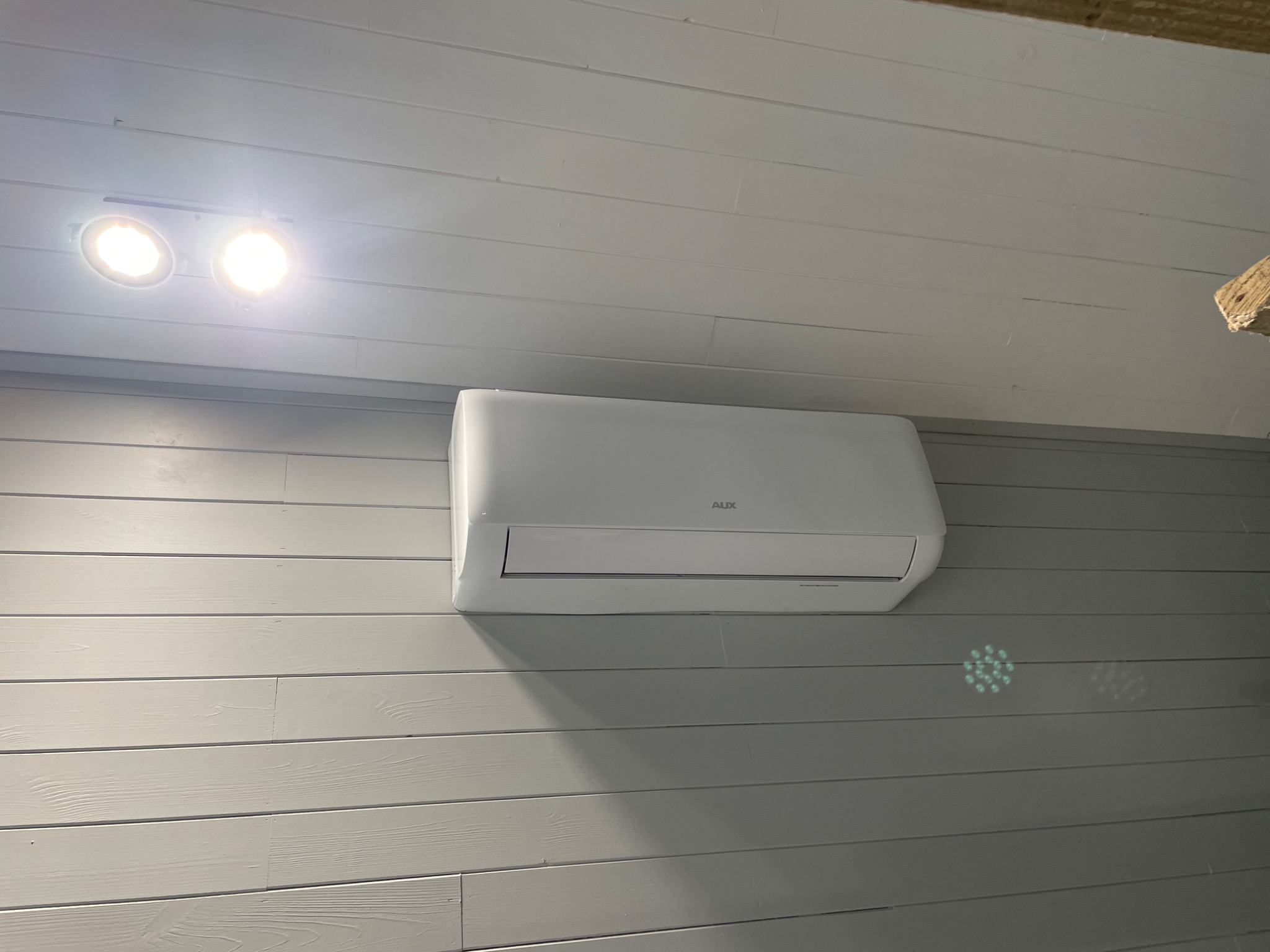Biała klimatyzacja AUX zamontowana na szarej, drewnianej ścianie z widocznym oświetleniem sufitowym i dekoracyjnym wzorem świetlnym na ścianie.