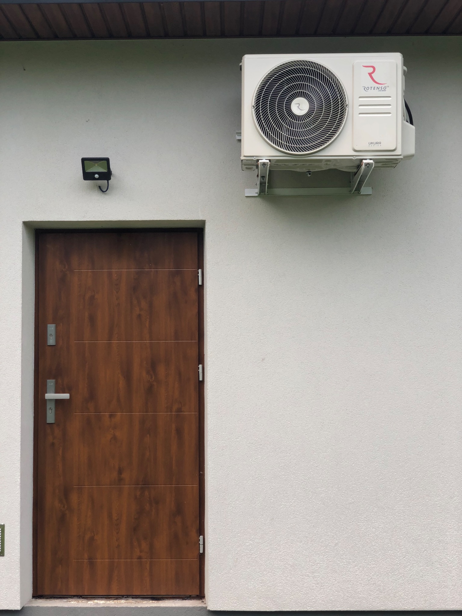 Zewnętrzna jednostka klimatyzacji Rotenso zamontowana na białej elewacji domu obok brązowych drzwi wejściowych z oświetleniem LED nad nimi.