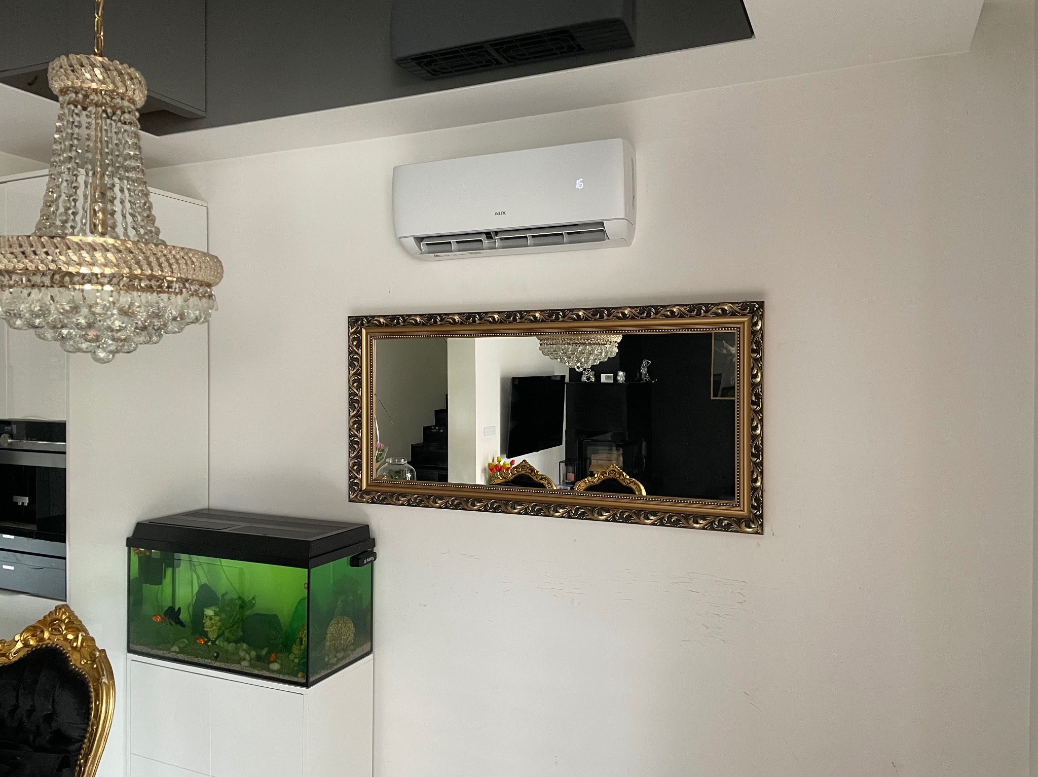Biały klimatyzator AUX zamontowany na ścianie w salonie, widoczny także kryształowy żyrandol, akwarium z rybkami i lustro w złotej ramie.