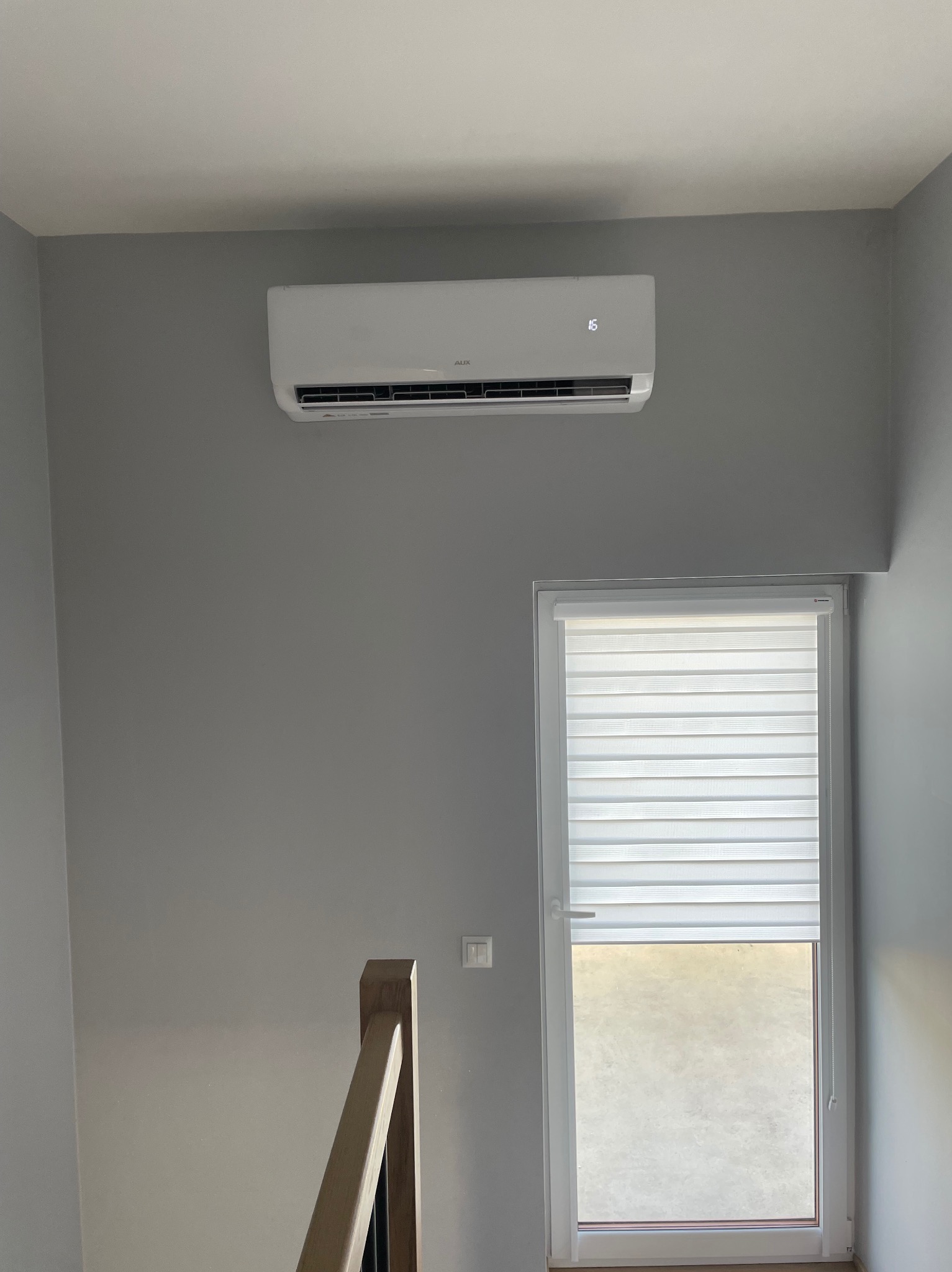 Biały klimatyzator ścienny AUX zamontowany w szarym pomieszczeniu nad oknem z żaluzjami, widok z dołu ze schodów.