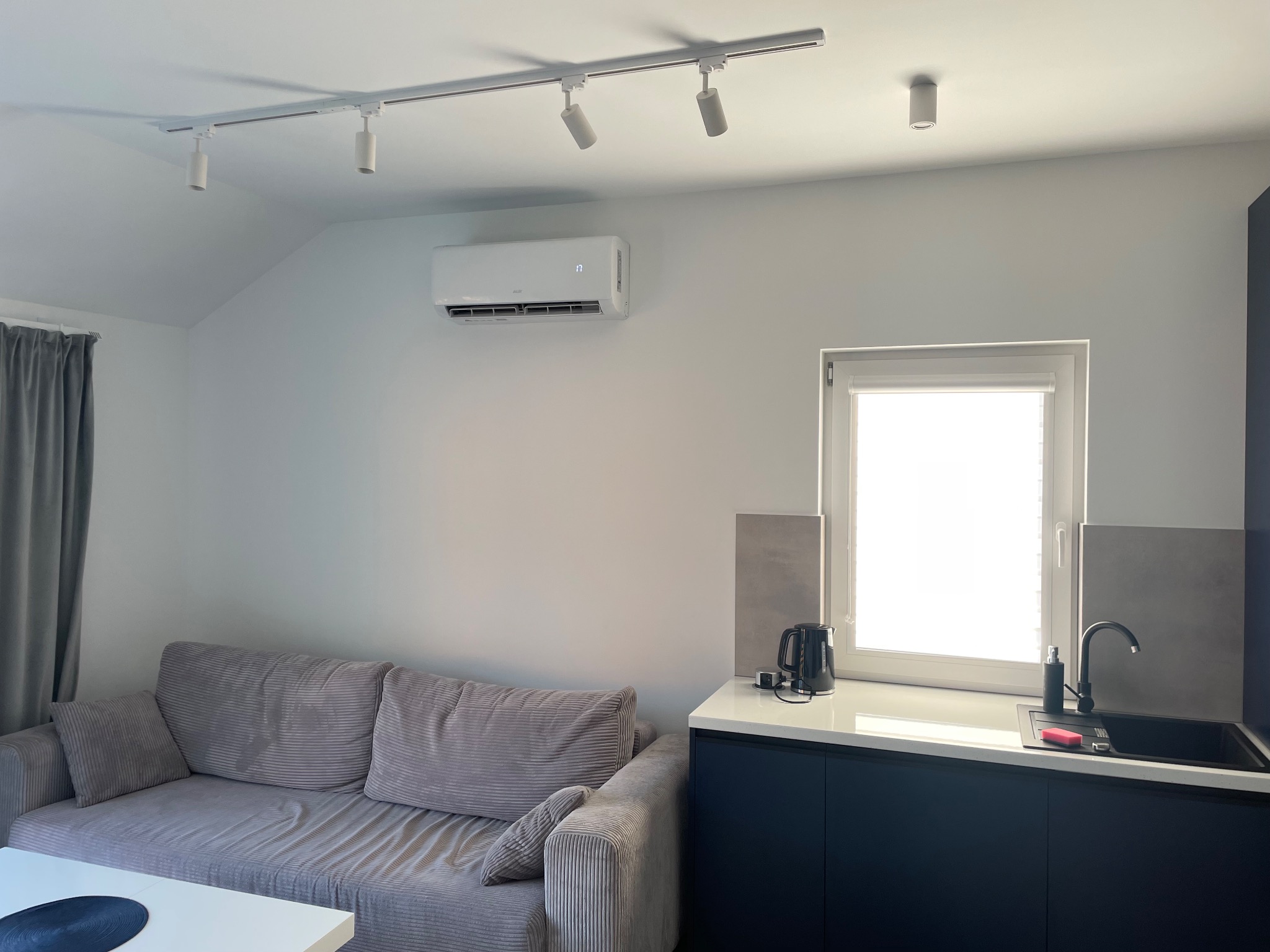 Wnętrze pokoju z klimatyzatorem zamontowanym na ścianie nad szarą sofą, widoczna również kuchnia z czarnym zlewem i czajnikiem, oświetlenie sufitowe punktowe.