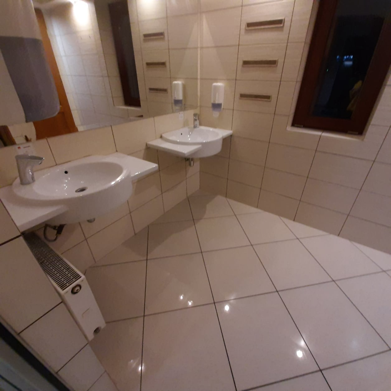 Umywalki w łazience z beżowymi płytkami, widoczny grzejnik i dozowniki mydła, odbicie w lustrze