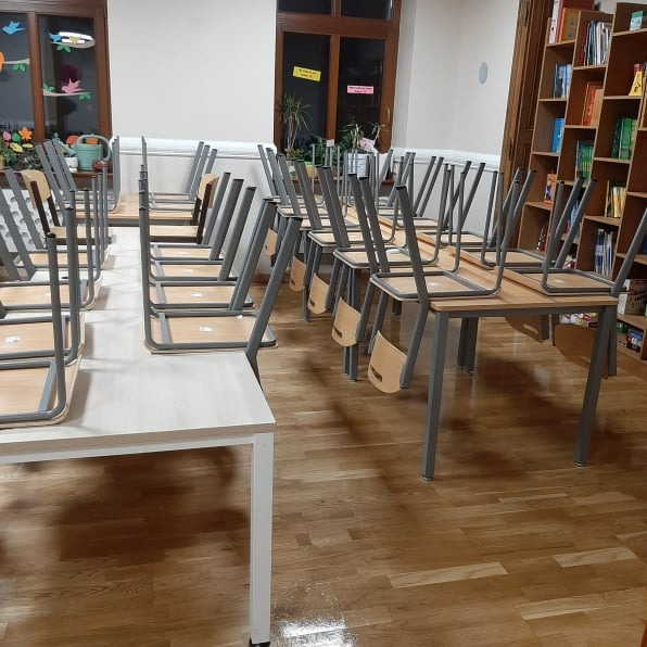 Ustawione na biurkach krzesła w pustej sali lekcyjnej po sprzątaniu, widoczne regały z książkami i dekoracje okienne.