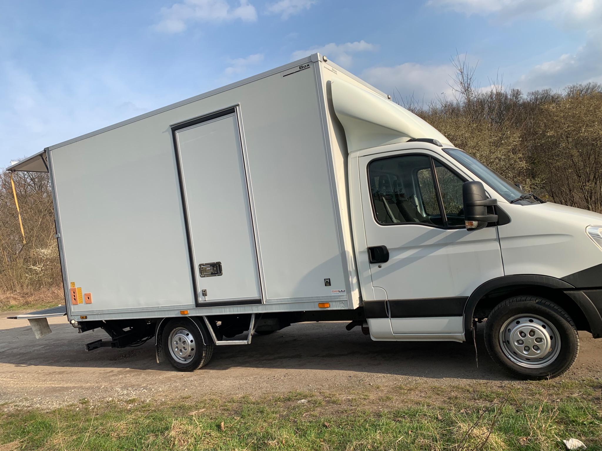 Biały samochód dostawczy z otwartą rampą załadunkową na szutrowej drodze, w tle drzewa i błękitne niebo.