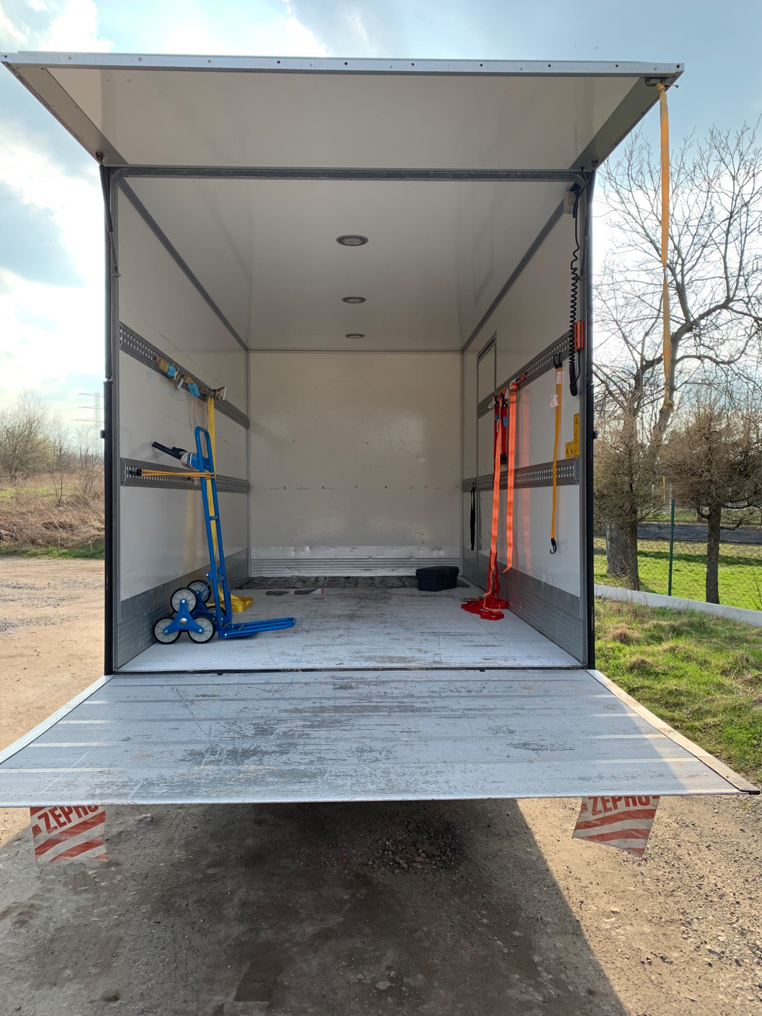 Wnętrze pustej, otwartej paki dostawczego samochodu z niebieskim wózkiem schodowym i pasami mocującymi widocznymi na ścianach, oświetlenie sufitowe, zarys drzew na zewnątrz.