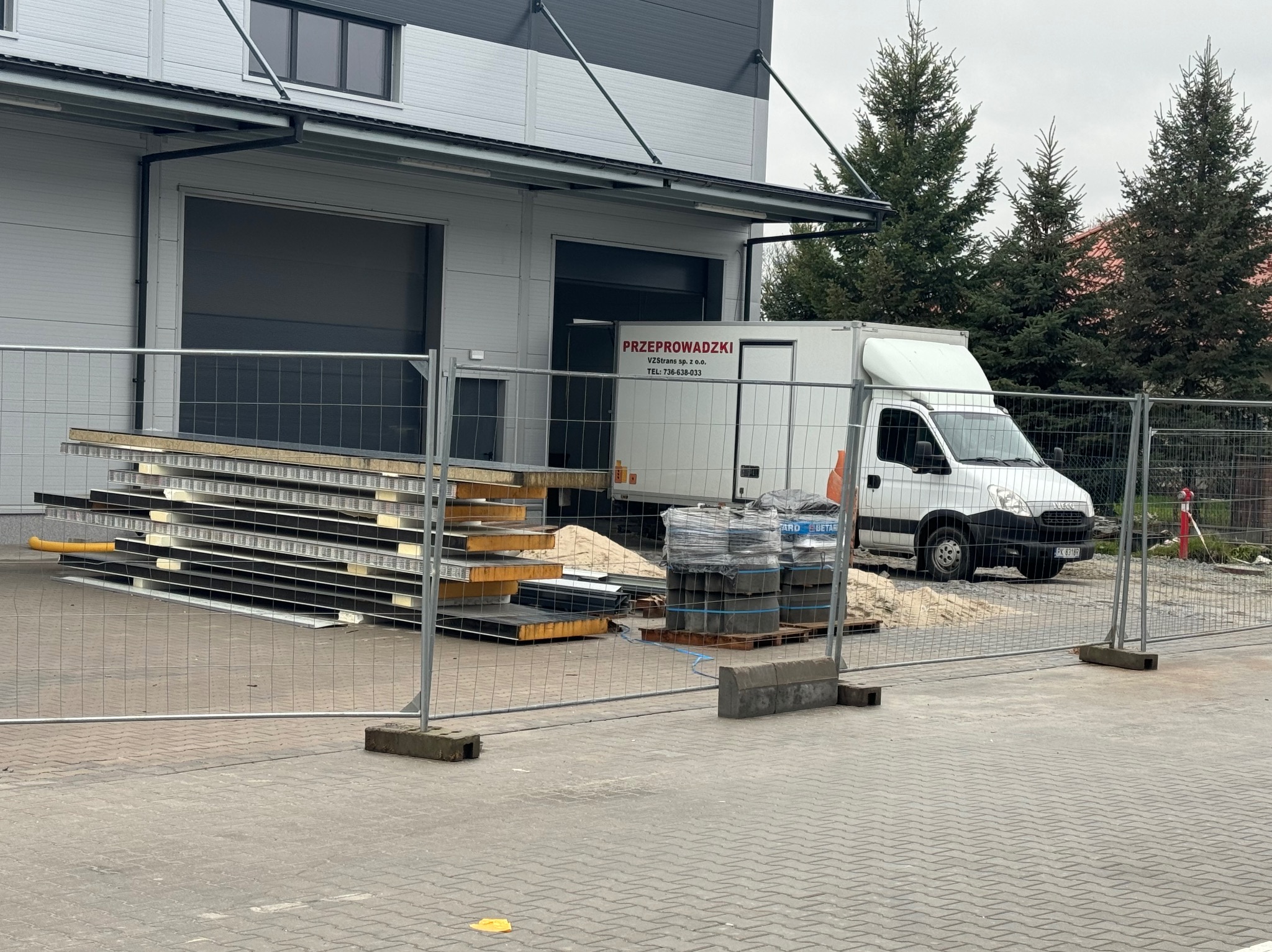 Biała furgonetka z napisem 'Przeprowadzki' zaparkowana przed magazynem z ładunkiem płyt warstwowych, widoczna przez ogrodzenie.