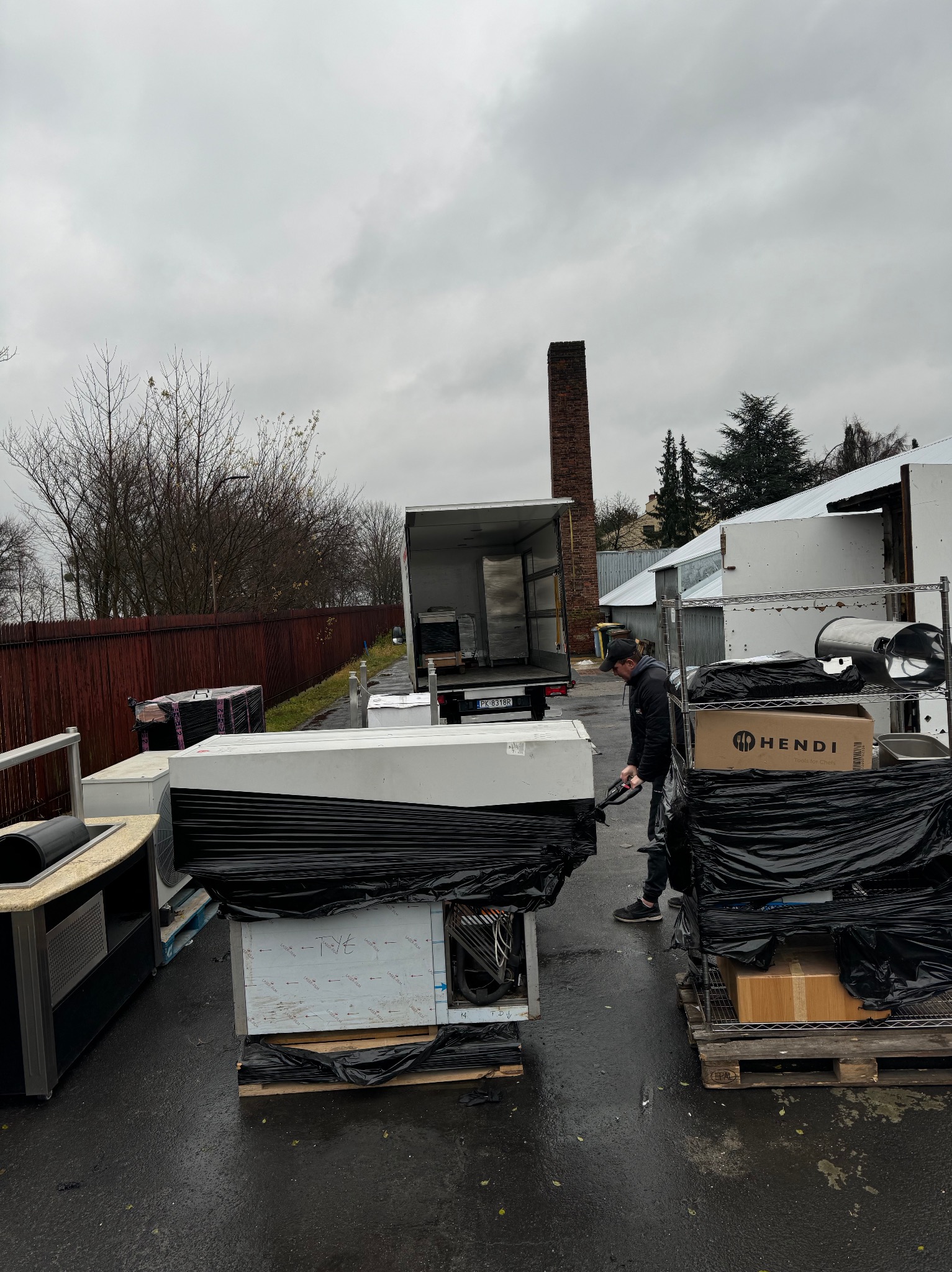Mężczyzna w czarnej kurtce i czapce rozładowuje z ciężarówki sprzęt gastronomiczny, w tle ceglany komin i szare niebo.