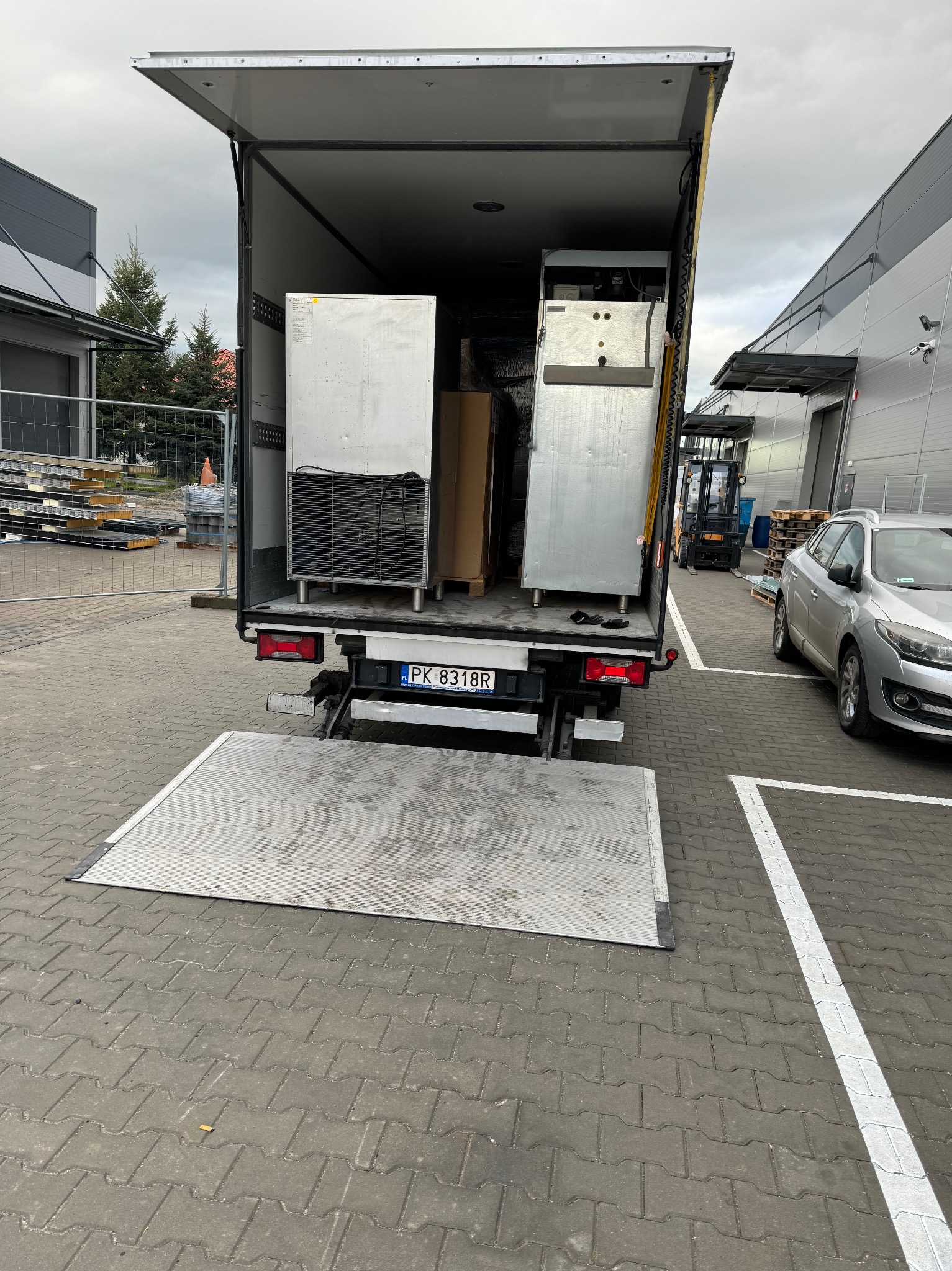 Otwarta paka busa z rampą załadowczą, wewnątrz dwie duże, srebrne chłodziarki przemysłowe zabezpieczone kartonami, widoczny wózek widłowy i zaparkowany samochód osobowy.