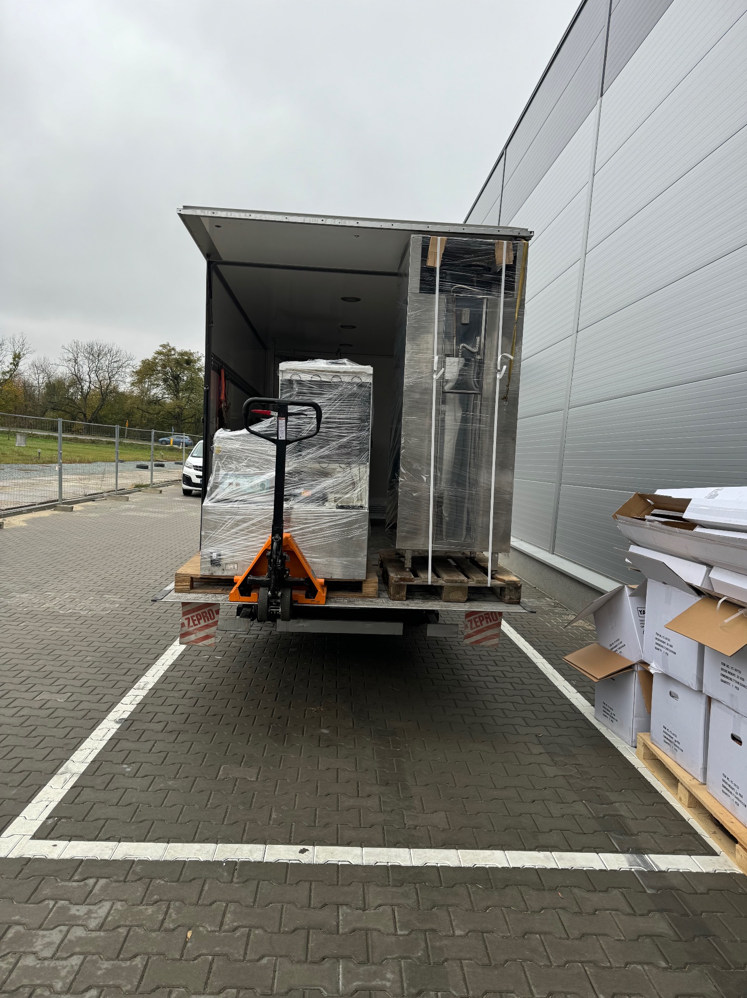 Załadunek palet z towarem owiniętym folią stretch do otwartego busa przy użyciu wózka paletowego, na parkingu z kostki brukowej, widoczne kartony obok.