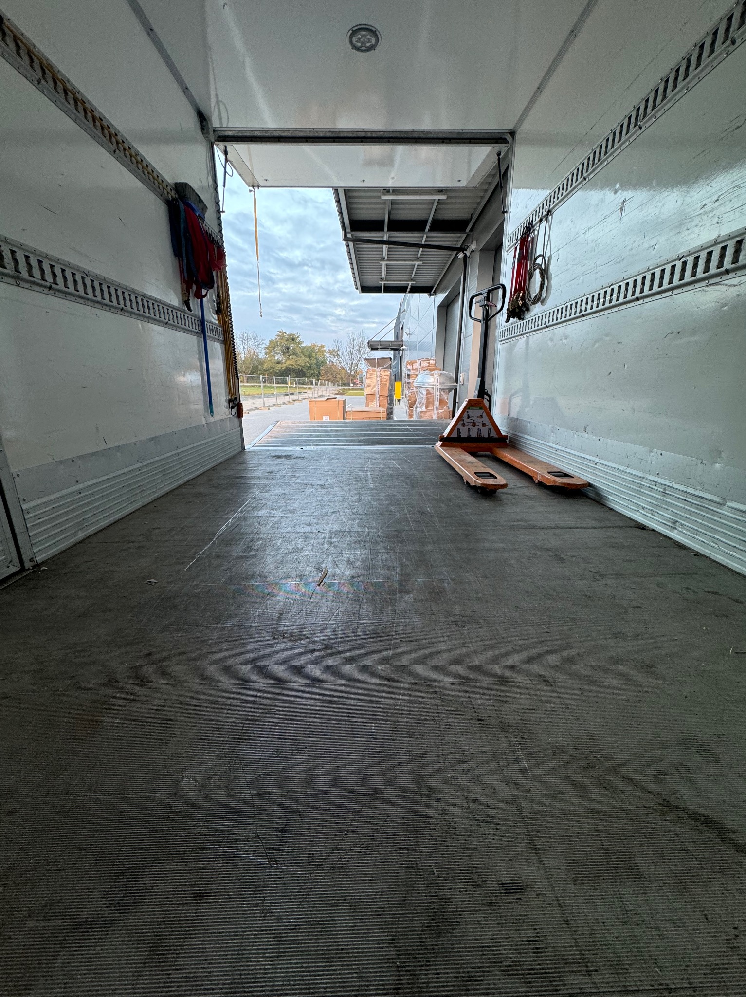 Wnętrze pustej przestrzeni ładunkowej pojazdu z wózkiem paletowym, widok na rampę załadunkową i plac z kostką brukową w pochmurny dzień.