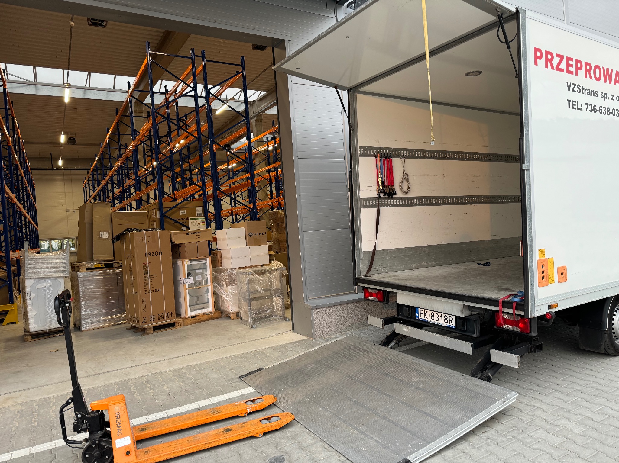 Wnętrze paki białego samochodu dostawczego z otwartą rampą załadunkową, pomarańczowy wózek paletowy przed rampą, w tle regały magazynowe z zapakowanymi kartonami, widoczna tablica rejestracyjna...