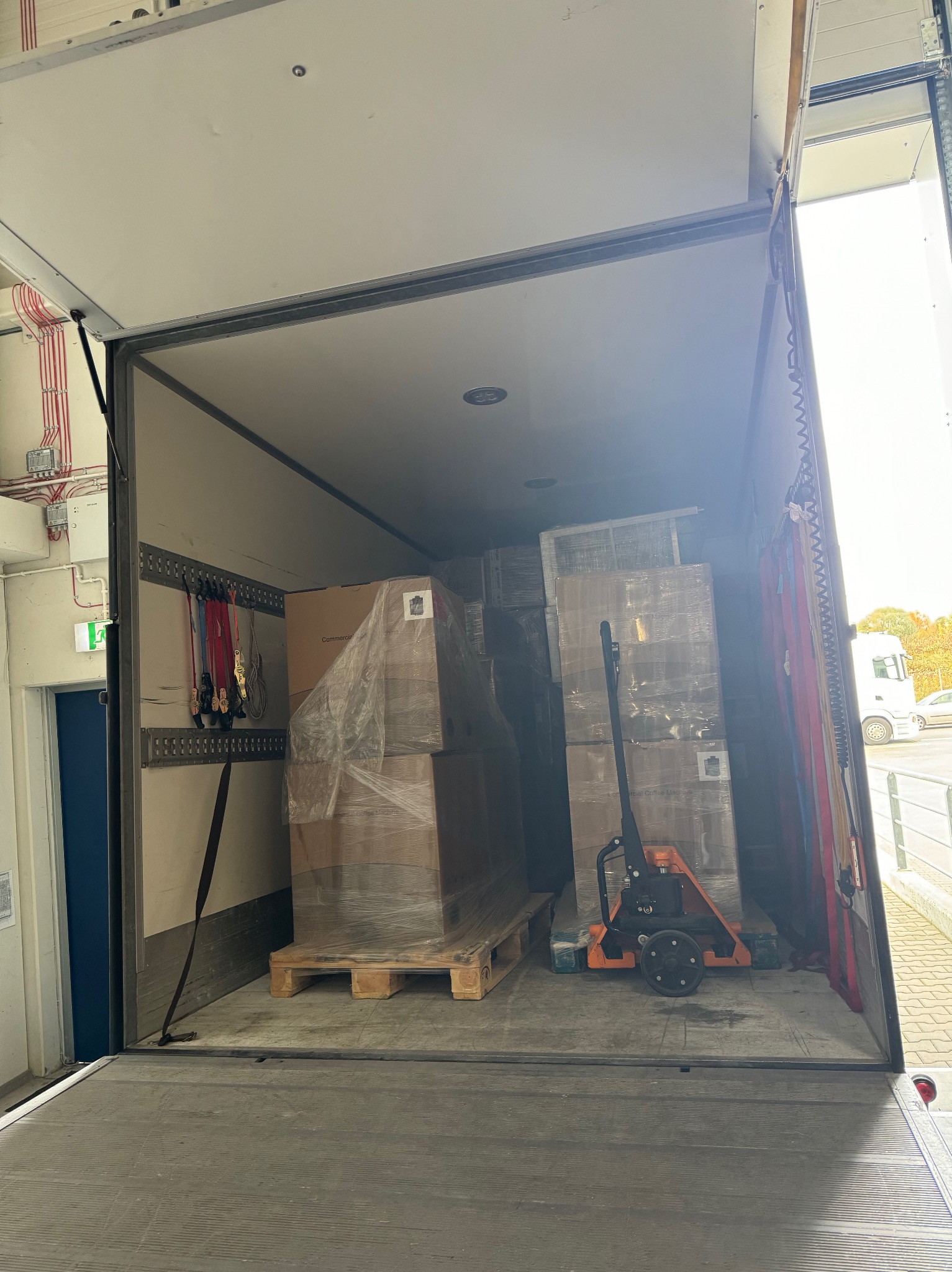 Wnętrze przestrzeni ładunkowej busa z paletami opakowanymi folią stretch i wózkiem paletowym; widoczne pasy mocujące ładunek na ścianie. Z zewnątrz widoczny fragment parkingu i zaparkowanego auta.