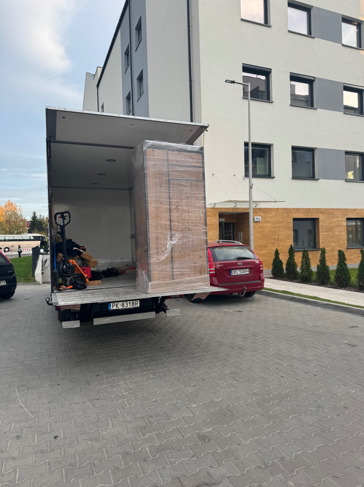 Mebel zabezpieczony folią stretch w otwartym samochodzie dostawczym z podnośnikiem ręcznym, zaparkowanym na kostce brukowej przed nowoczesnym budynkiem mieszkalnym.