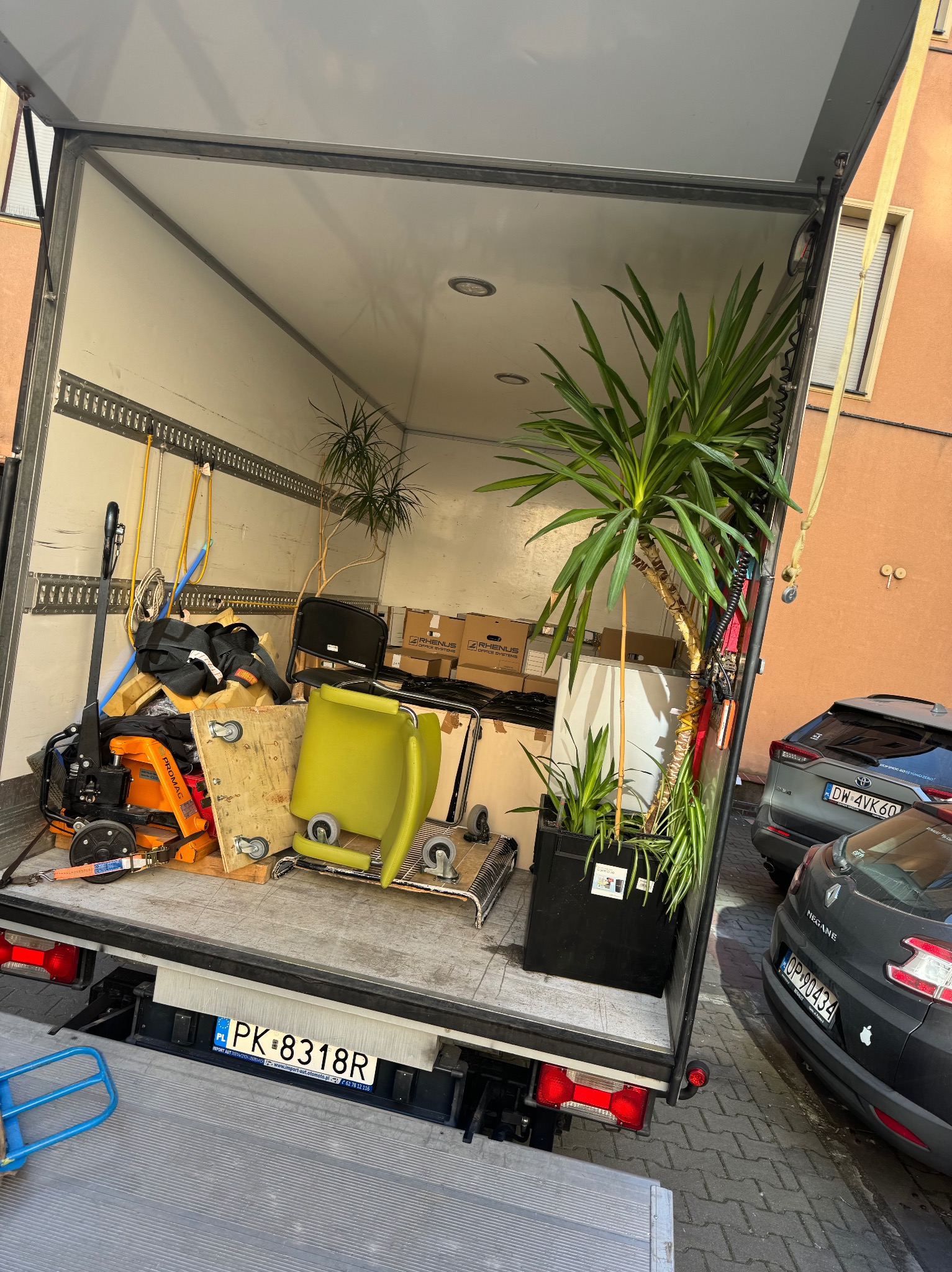 Wnętrze furgonetki załadowanej meblami biurowymi, roślinami doniczkowymi i sprzętem do transportu, widoczne tablice rejestracyjne pojazdów na zewnątrz