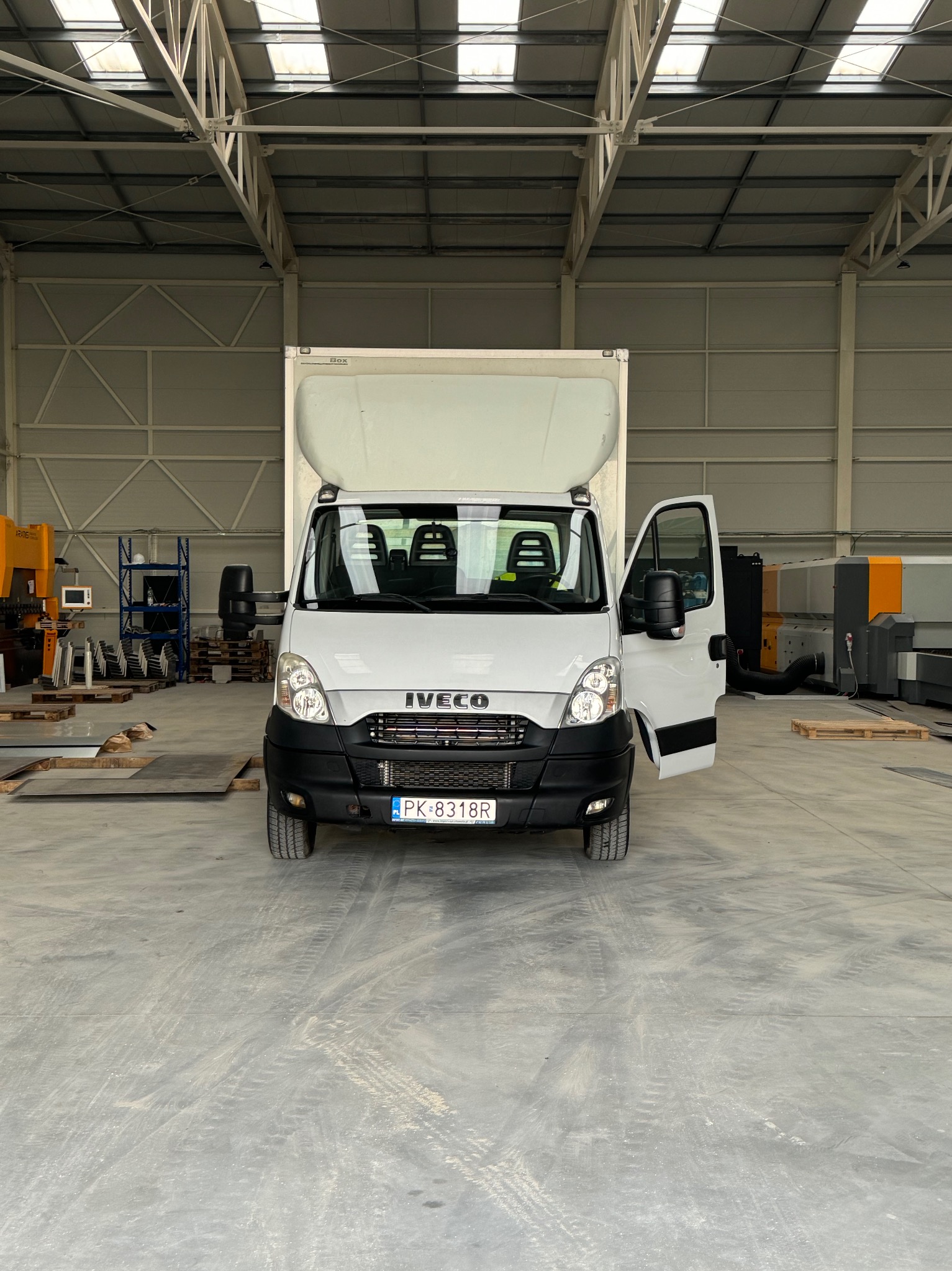 Biały samochód dostawczy Iveco z otwartymi drzwiami pasażera, zaparkowany wewnątrz hali przemysłowej z widocznymi maszynami i regałami w tle.