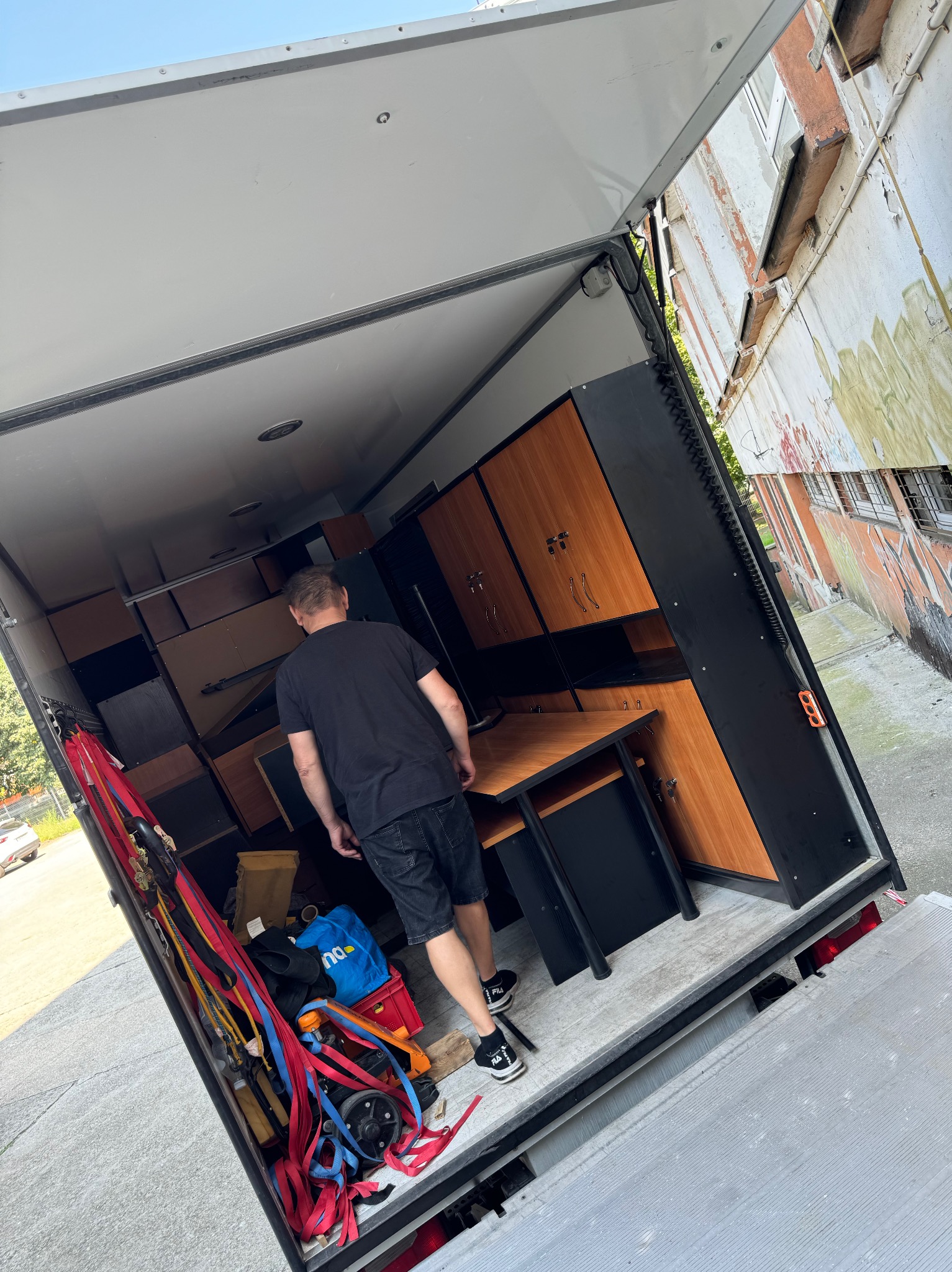 Mężczyzna w czarnej koszulce wchodzi do wnętrza furgonetki załadowanej meblami biurowymi, w tle widoczny budynek z graffiti.