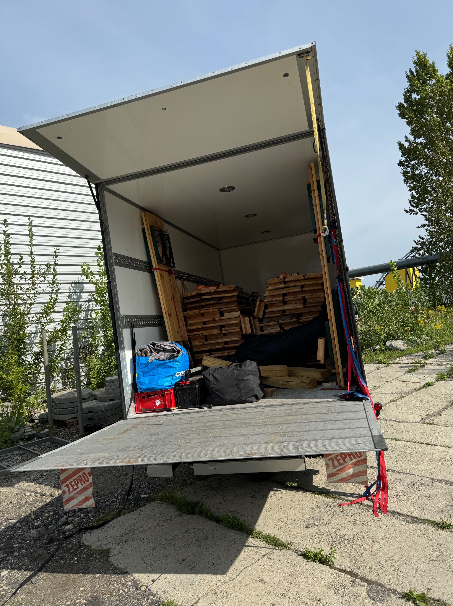 Otwarty tył furgonetki załadowany paletami, drewnianymi deskami i workami, zabezpieczony pasami transportowymi, widok z zewnątrz na betonowy plac i zieleń