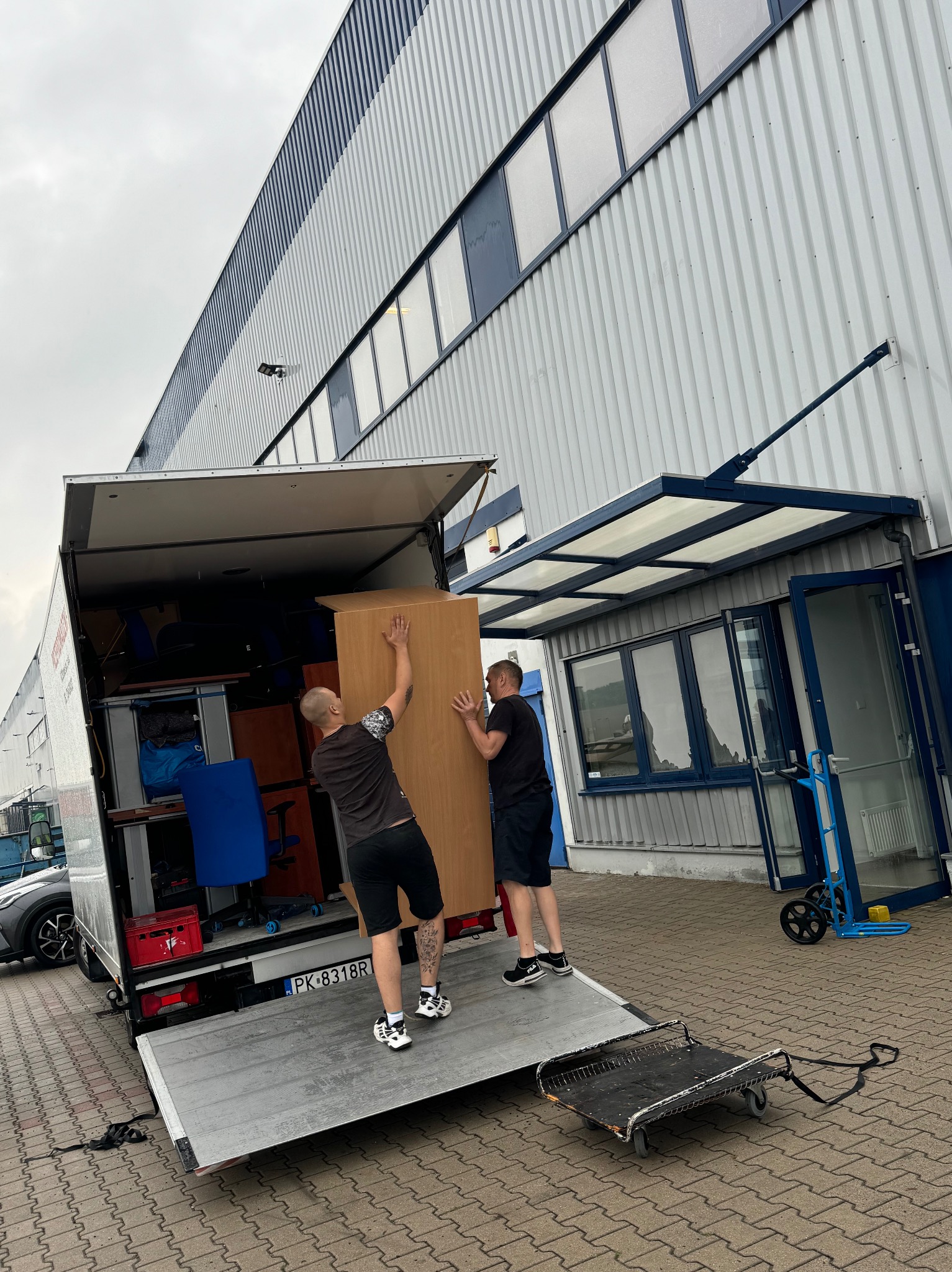 Dwóch mężczyzn ładuje drewniany mebel do furgonetki podczas przeprowadzki we Wrocławiu, widoczny wózek transportowy i wnętrze wypełnione meblami.
