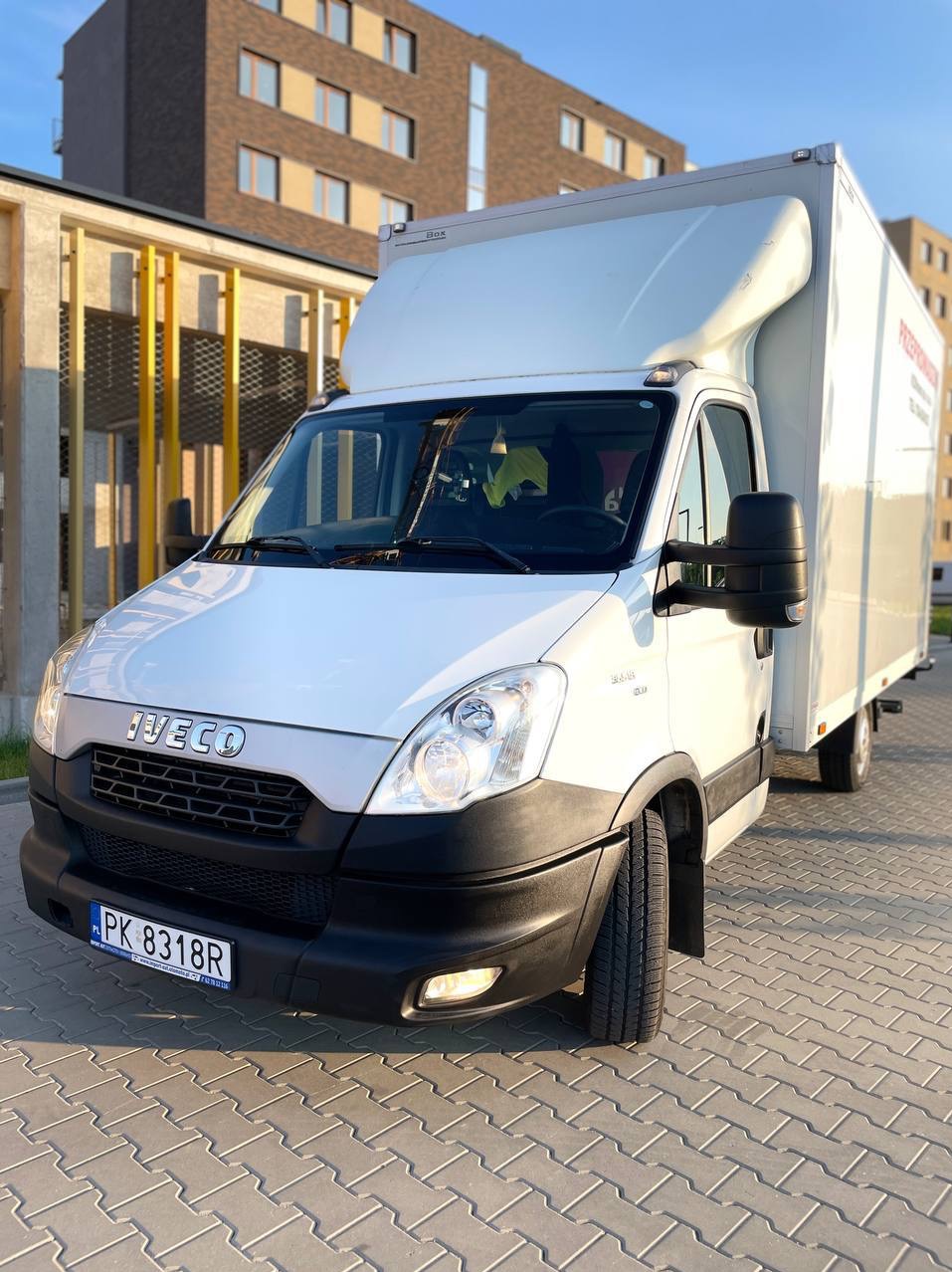 Biały samochód dostawczy Iveco z zabudową kontenerową, zaparkowany na brukowanym placu przed nowoczesnym budynkiem mieszkalnym w słoneczny dzień. Na boku widoczny napis.