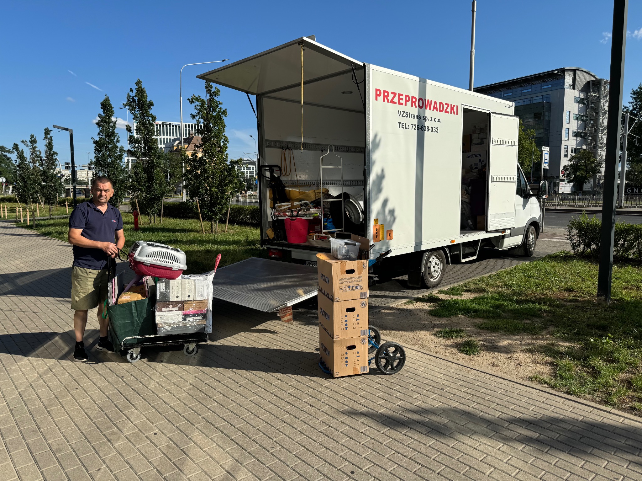 Mężczyzna z wózkiem pełnym rzeczy, w tym transporterem dla zwierząt, na tle otwartego busa z napisem 'PRZEPROWADZKI' i stertą kartonów na chodniku.
