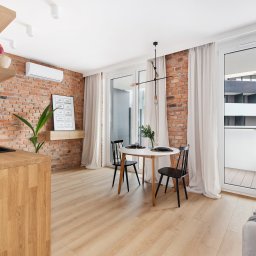 Krzysztof Błaszczyk PUH Booloo - Jasne wnętrze mieszkania z ceglaną ścianą, drewnianym blatem kuchennym i podłogą, okrągłym stołem z czarnymi krzesłami, balkonem oraz minimalistyczną lampą sufitową.