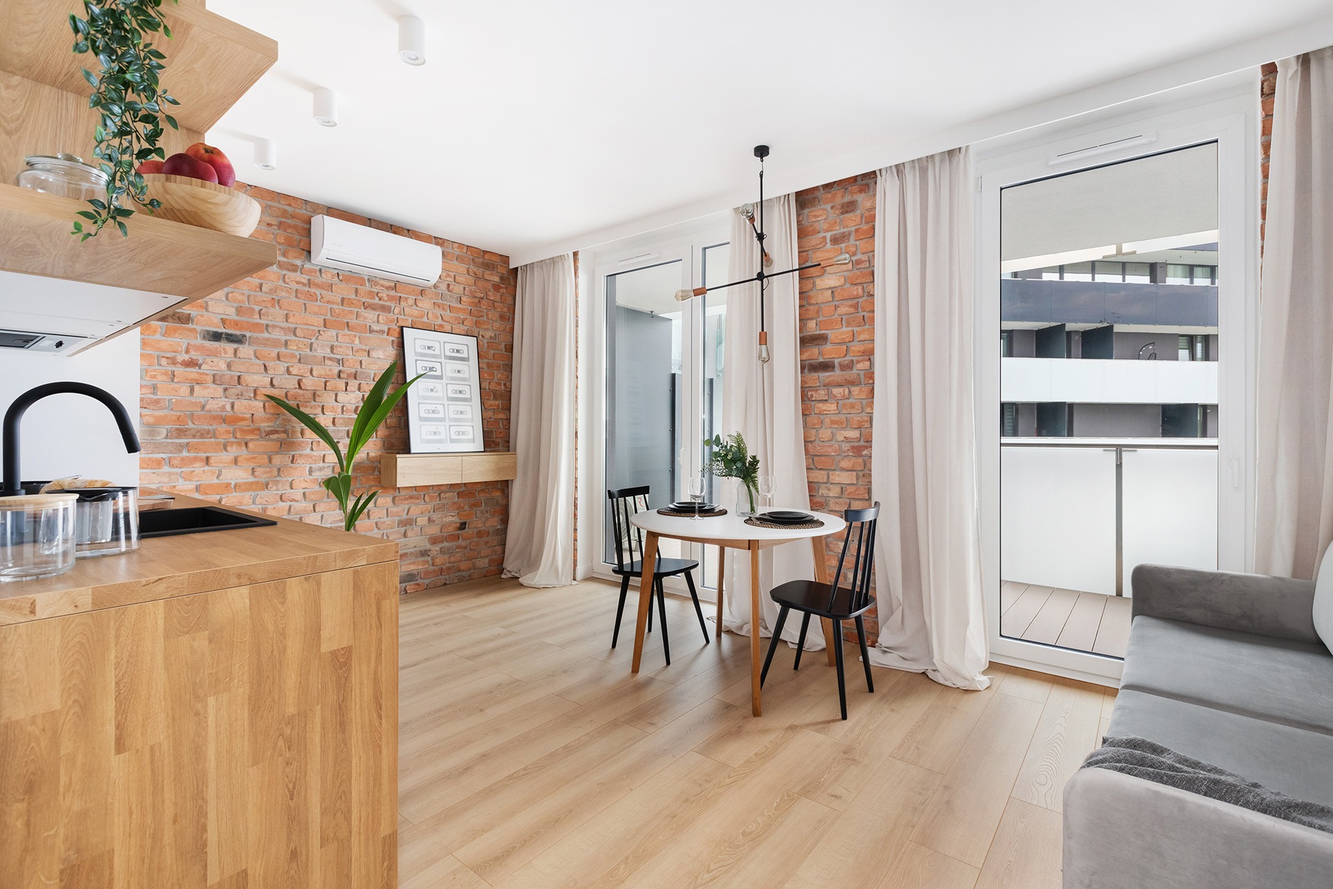 Jasne wnętrze mieszkania z ceglaną ścianą, drewnianym blatem kuchennym i podłogą, okrągłym stołem z czarnymi krzesłami, balkonem oraz minimalistyczną lampą sufitową.