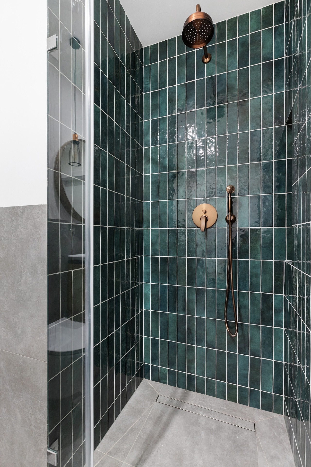 Kabina prysznicowa z ciemnozielonymi, pionowymi płytkami i miedzianą armaturą, widoczny odpływ liniowy w szarej posadzce.