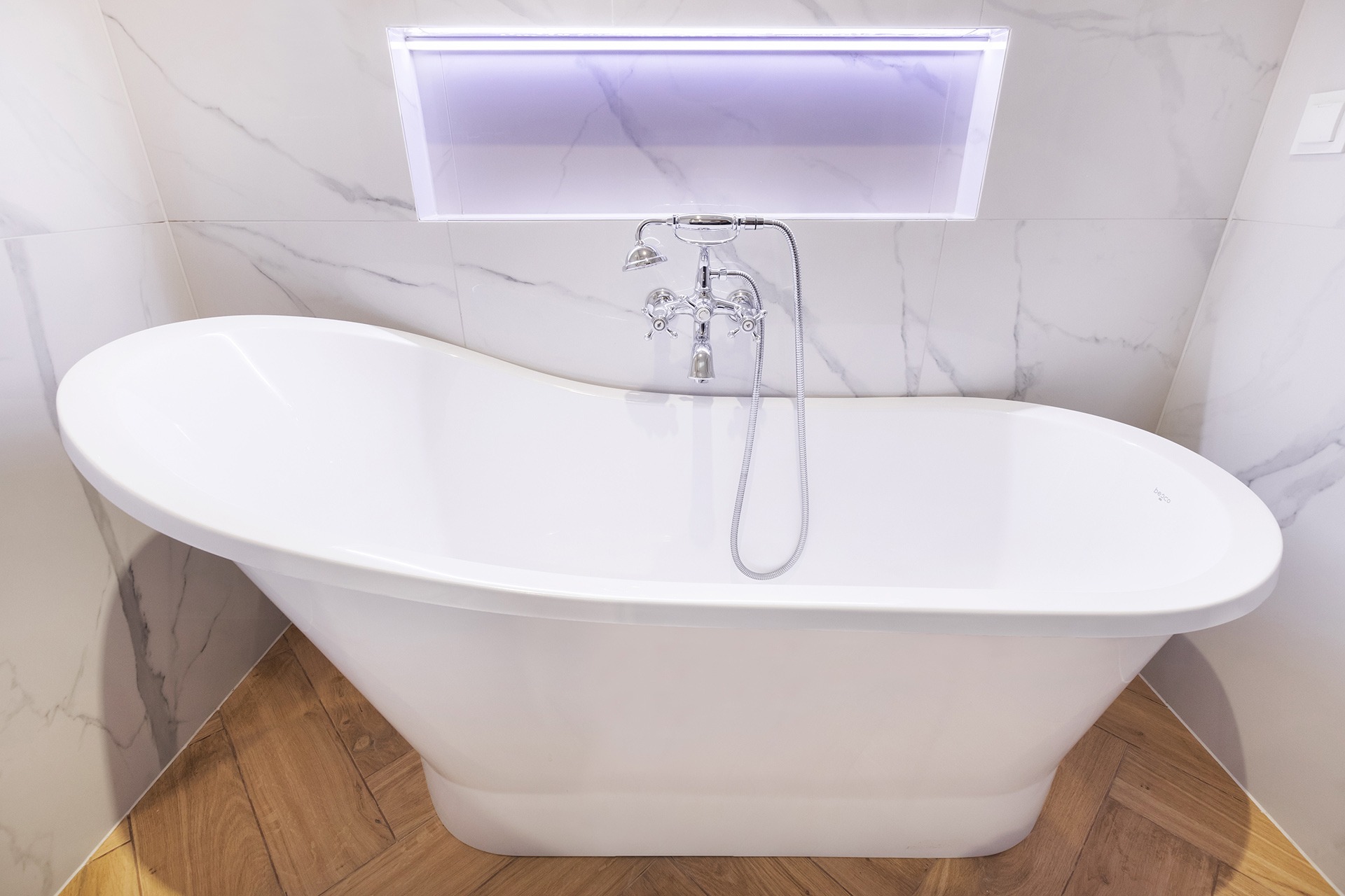 Wolnostojąca biała wanna z chromowaną armaturą na tle marmurowych płytek i drewnopodobnej podłogi ułożonej w jodełkę, oświetlenie LED w niszy nad wanną.